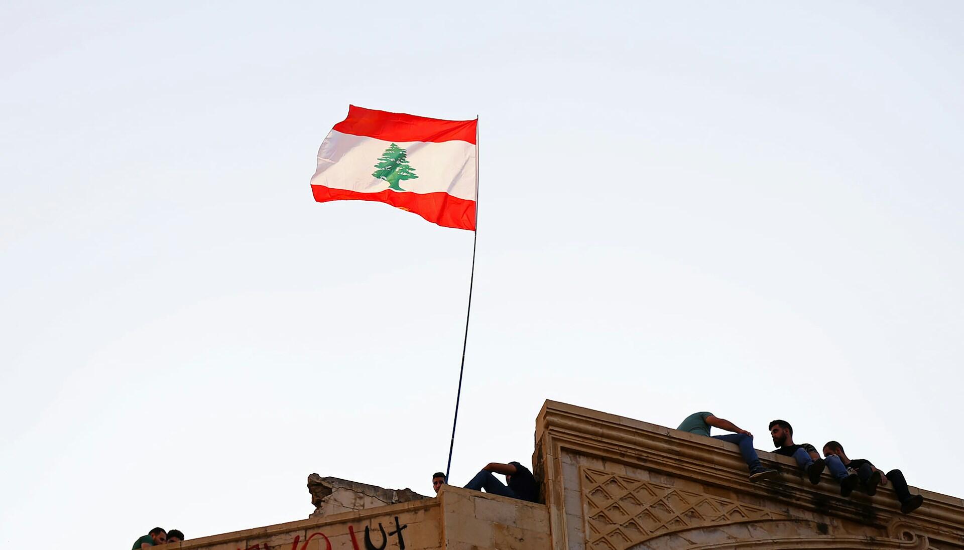 A Lebanese flag with a cedar tree design stands tall against the sky, while individuals sit on the ledge of a historic building.