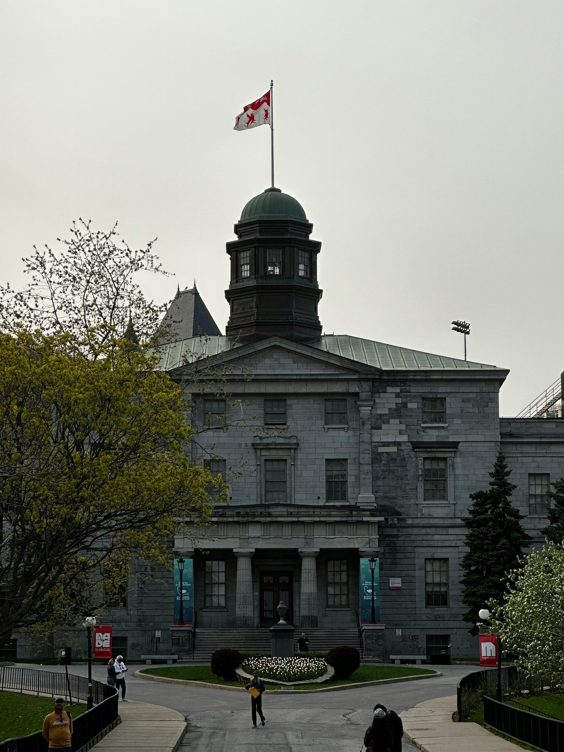 Historic stone building with a green domed roof, Canadian flag, surrounded by trees and blooming flowers, under an overcast sky.