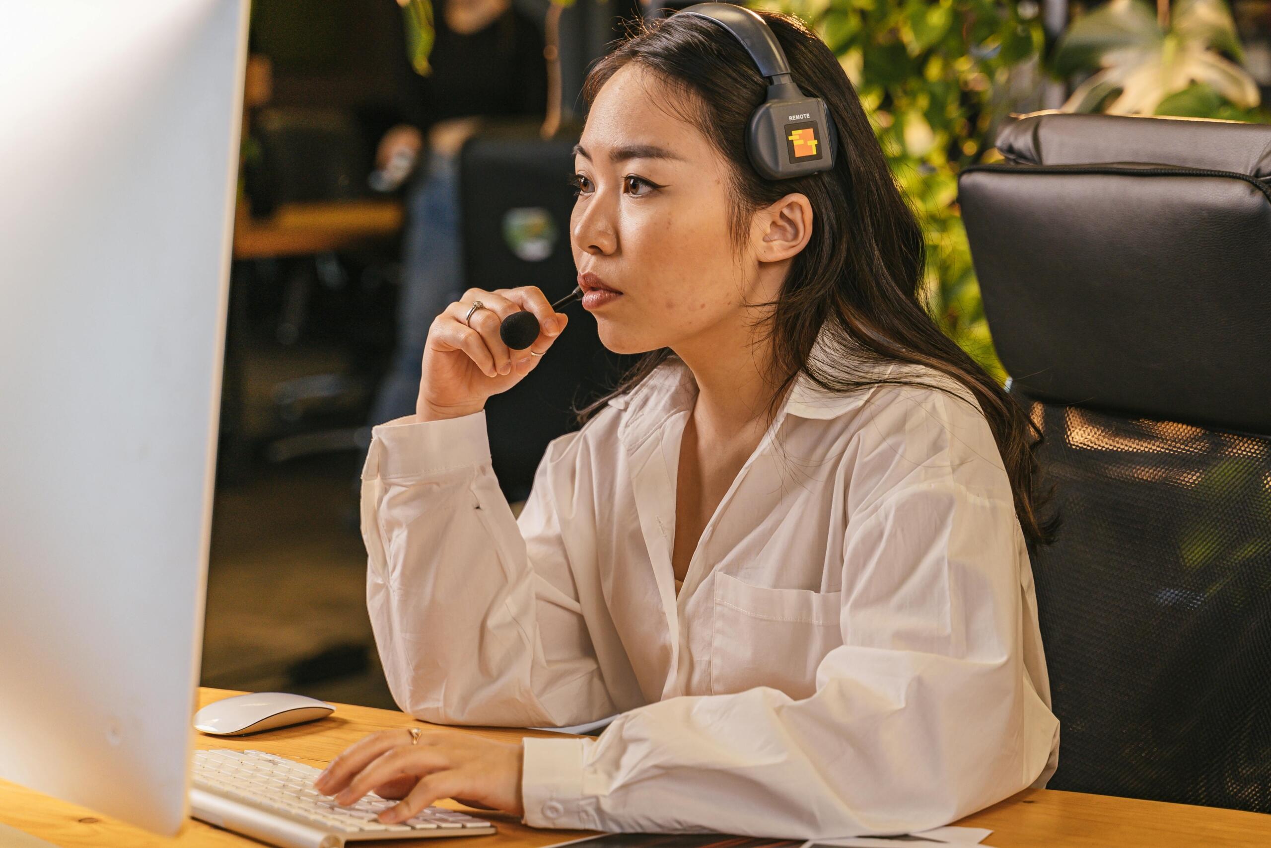 A woman wearing a white blouse and a headset, working at a computer, focused on a virtual meeting.