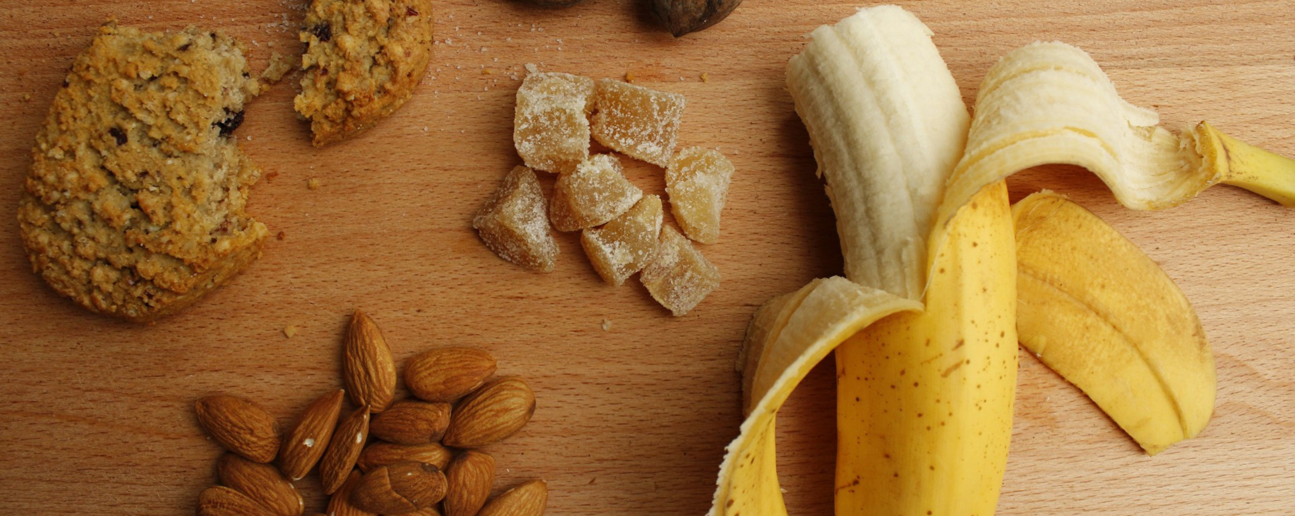 An assortment of snacks on a wooden surface: a cookie, almonds, candied ginger, and a peeled banana.