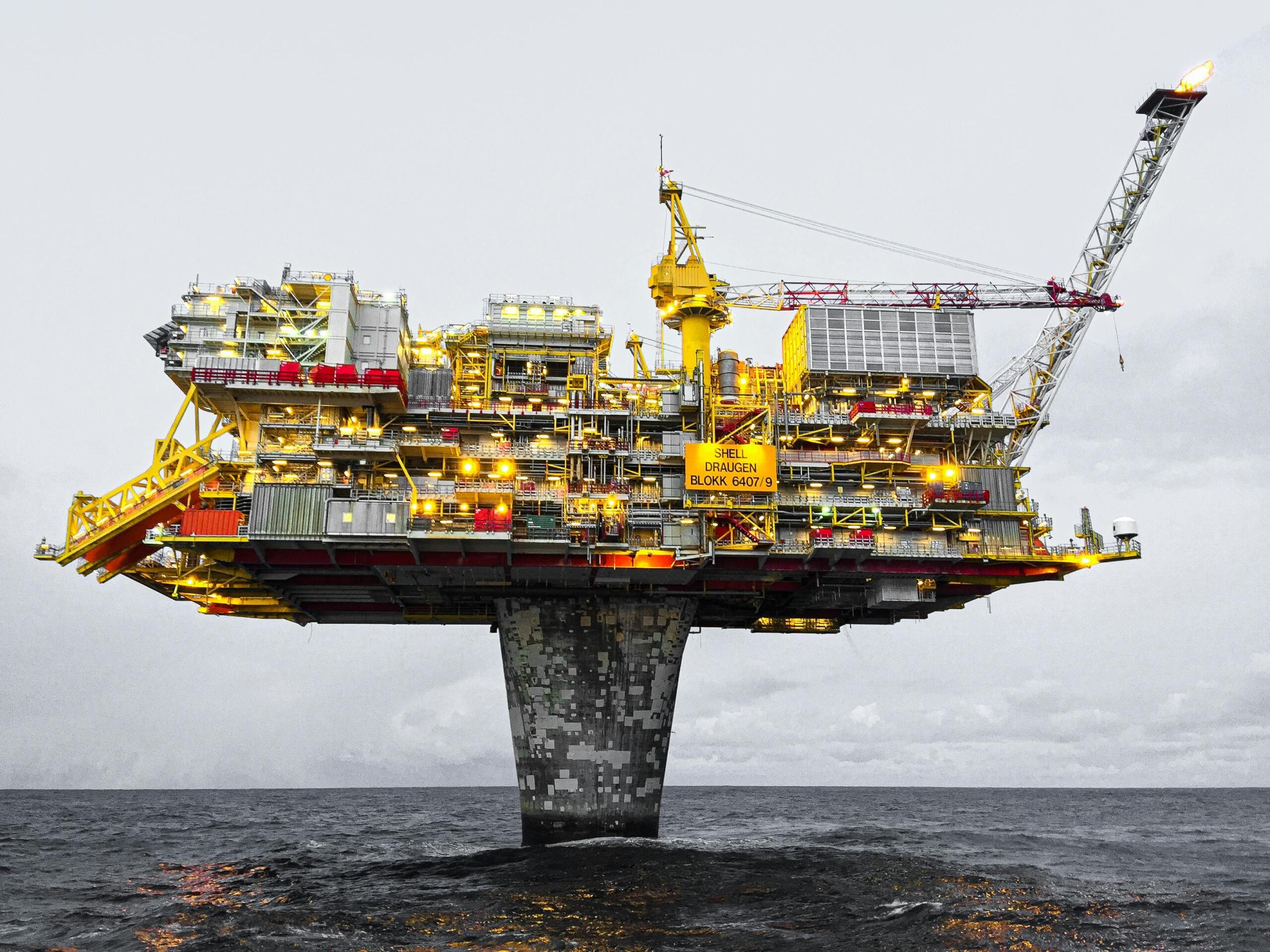 Large offshore oil platform standing in the ocean under grey skies, lit with yellow safety lights.