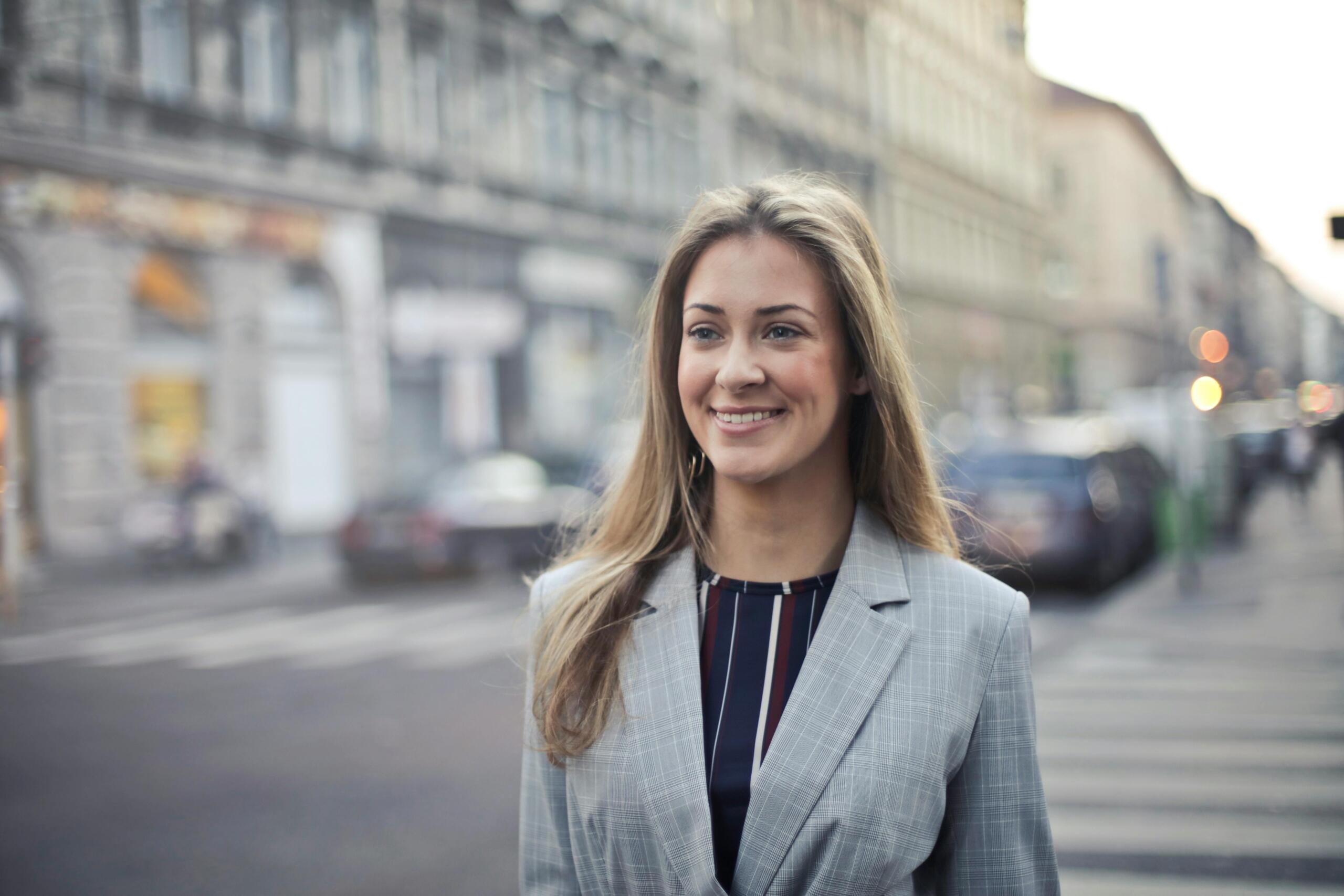 A woman in a blazer walks confidently through a bustling city street, surrounded by buildings and urban life.