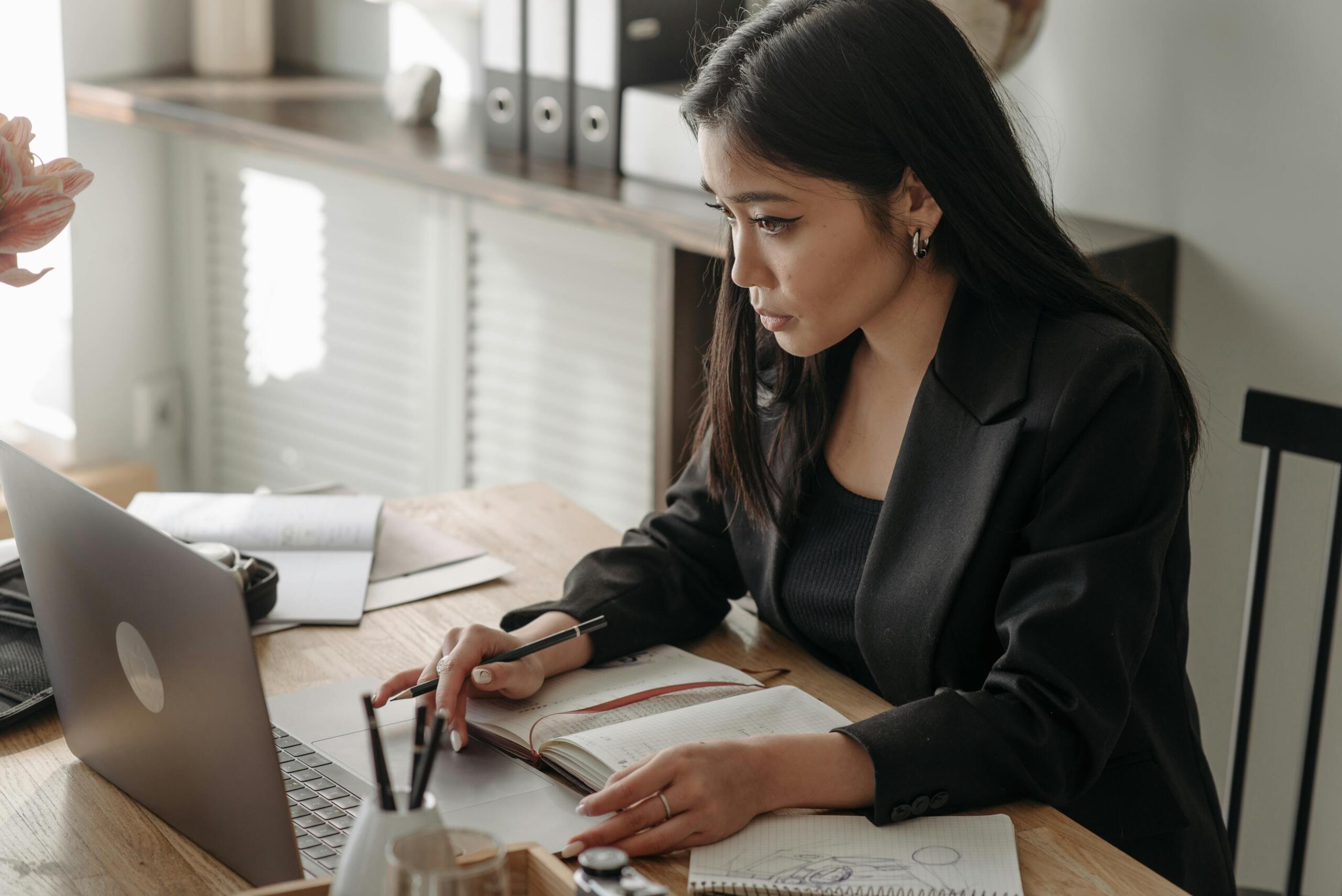 A woman in a business suit sits at a desk, focused on her laptop as she studies business courses online.