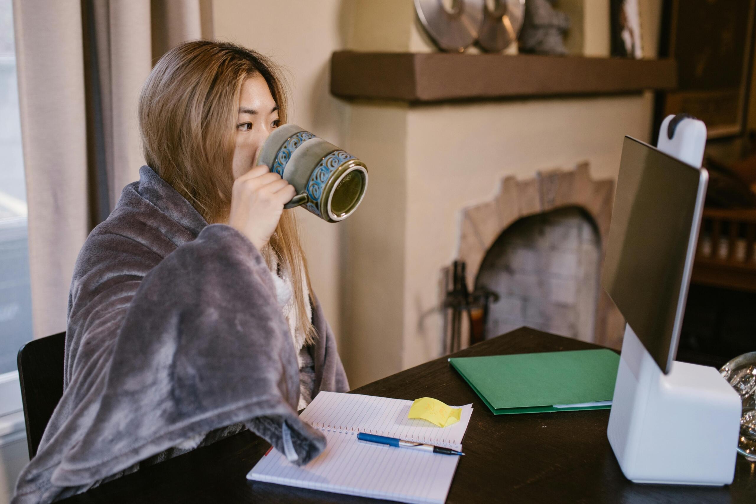A woman drinking coffee while studying online on her laptop, sitting comfortably at a table. She wears a blanket around her shoulders.