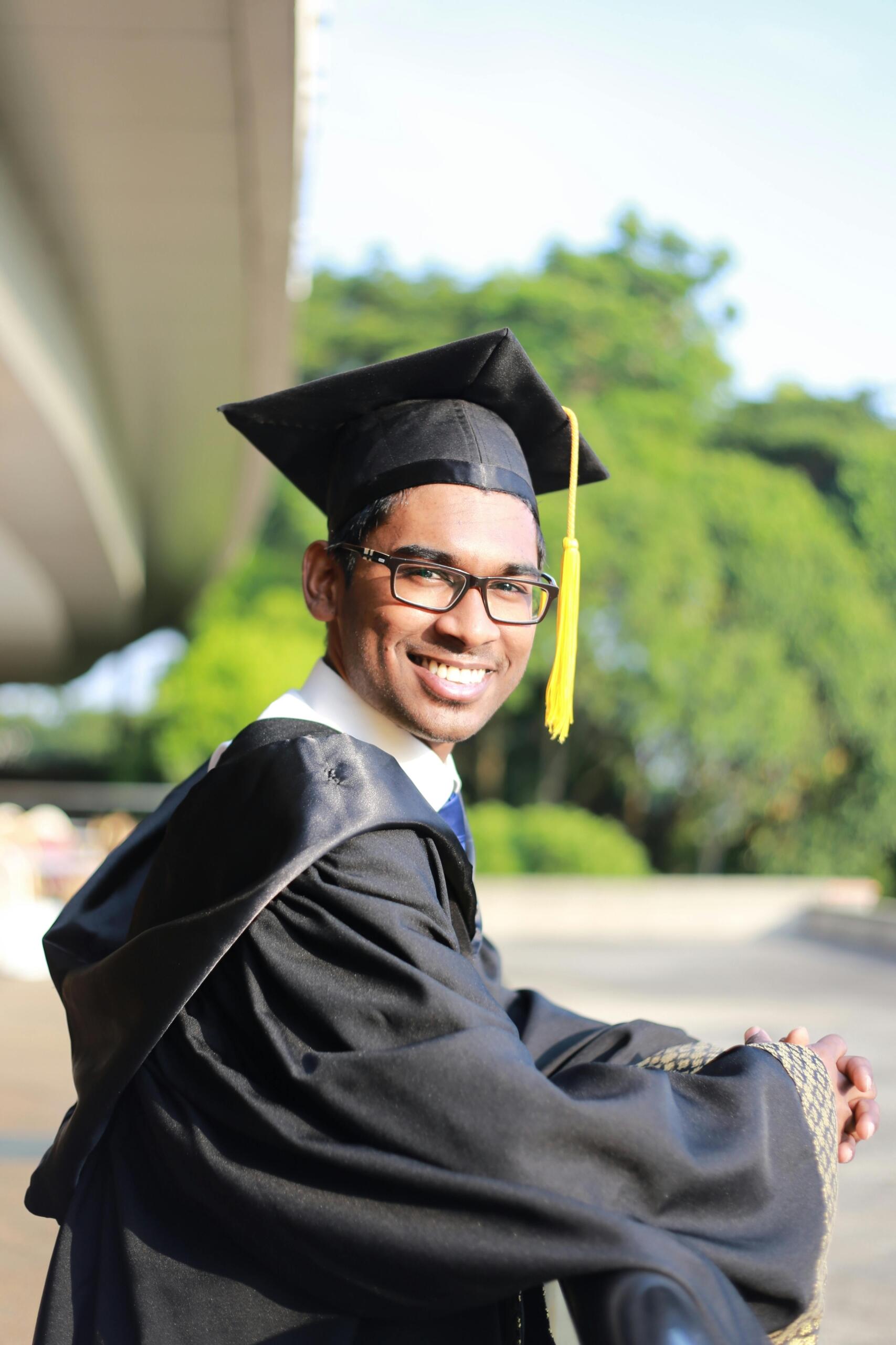A man wearing a cap and gown, smiling proudly in front of a backdrop of concrete and greenery. Source: Anant Raj.