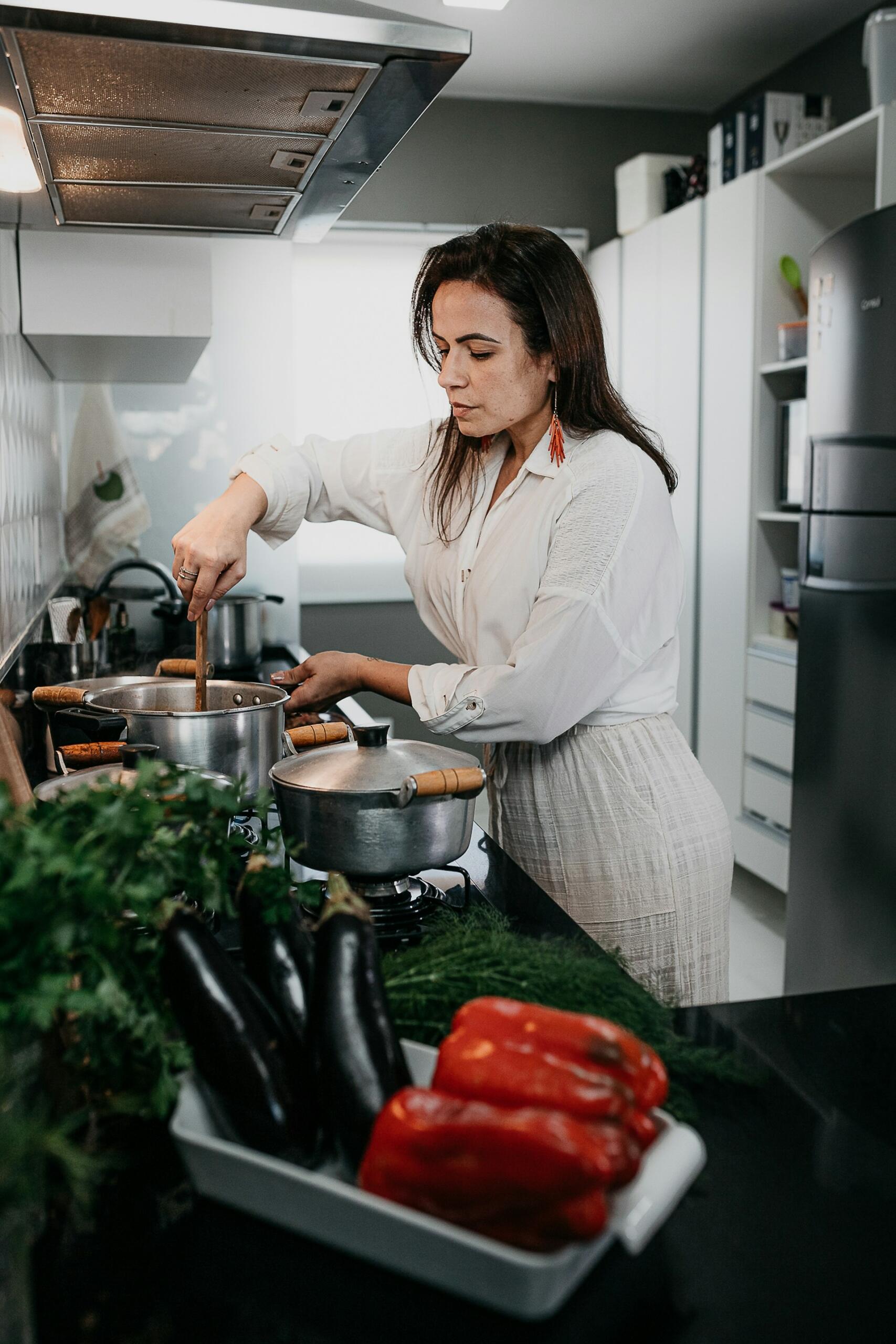 A woman stirs a pot in a modern kitchen, surrounded by fresh vegetables, including red peppers and eggplants, on the countertop.