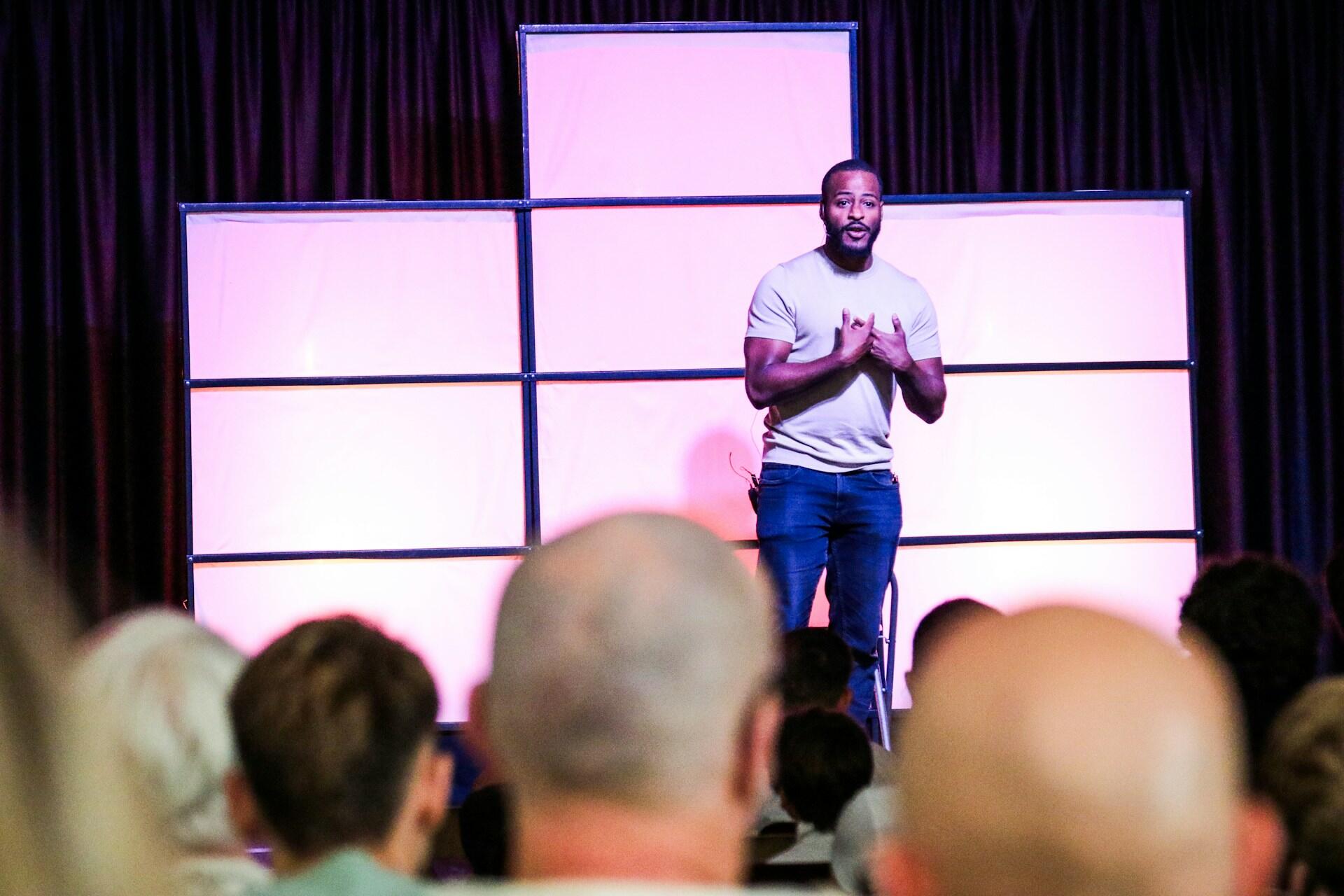 A speaker stands on stage with a purple backdrop, delivering a presentation to an attentive audience in a softly lit venue.