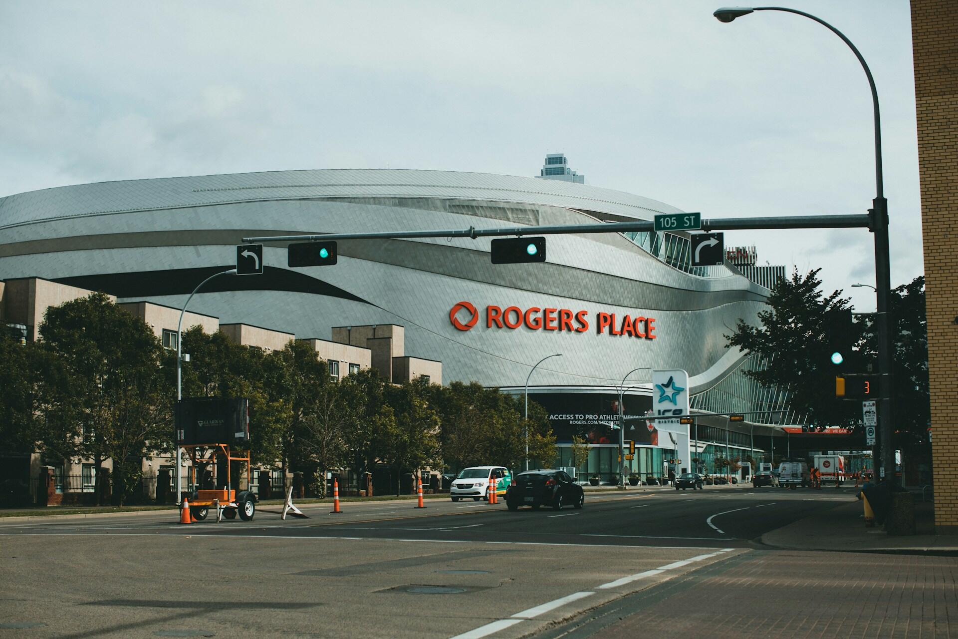 Rogers Place arena with a sleek silver exterior, featuring its name prominently, seen from a city street with traffic lights and trees.