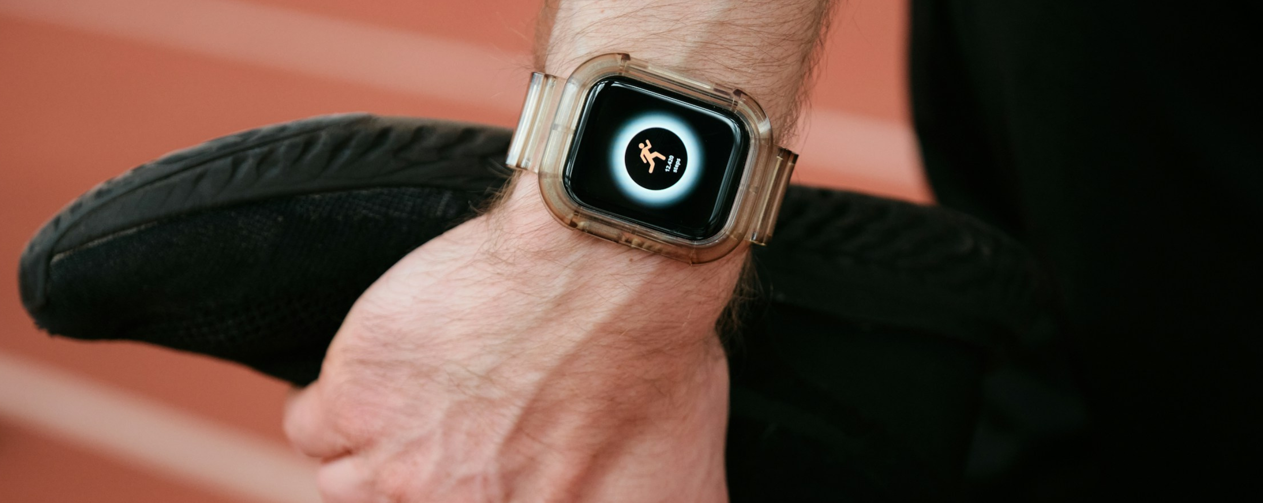 A close-up of a hand wearing a smartwatch displaying an icon of a running figure, set against a blurred athletic track background.