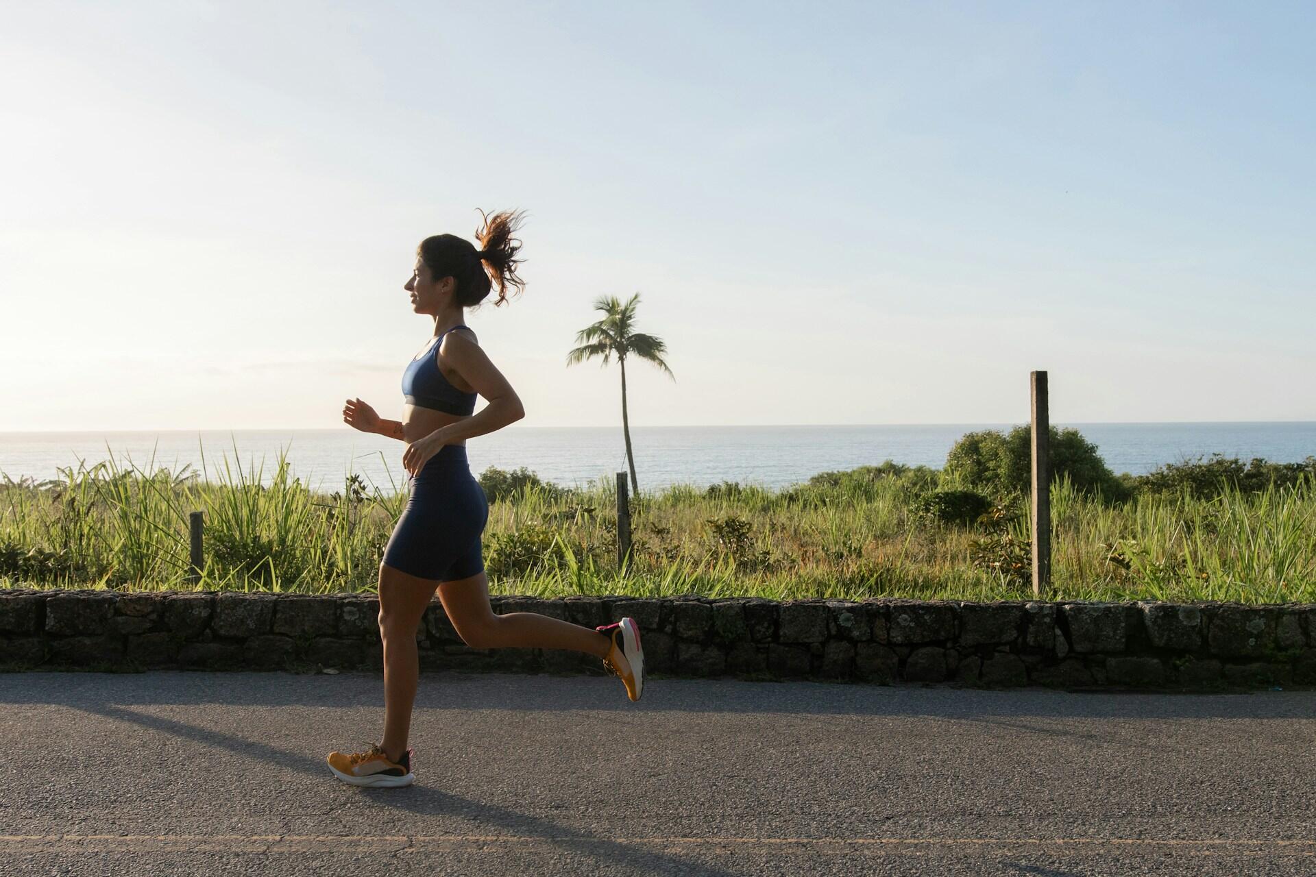A woman jogs along a coastal road, with lush grass and a serene ocean backdrop under a clear blue sky.