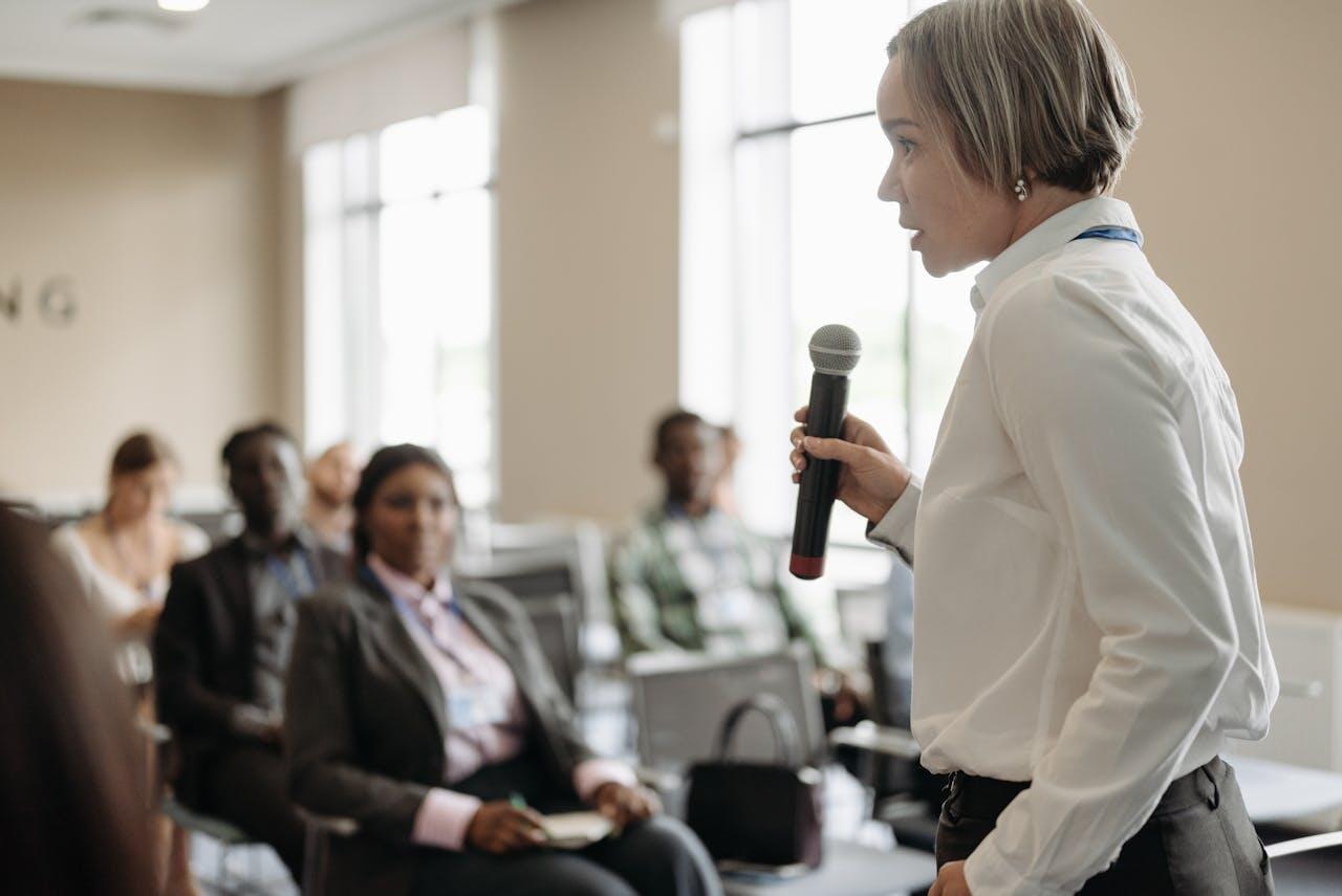 A speaker in a white blouse holds a microphone, engaging an audience in a professional setting with chairs lined up in a conference room.