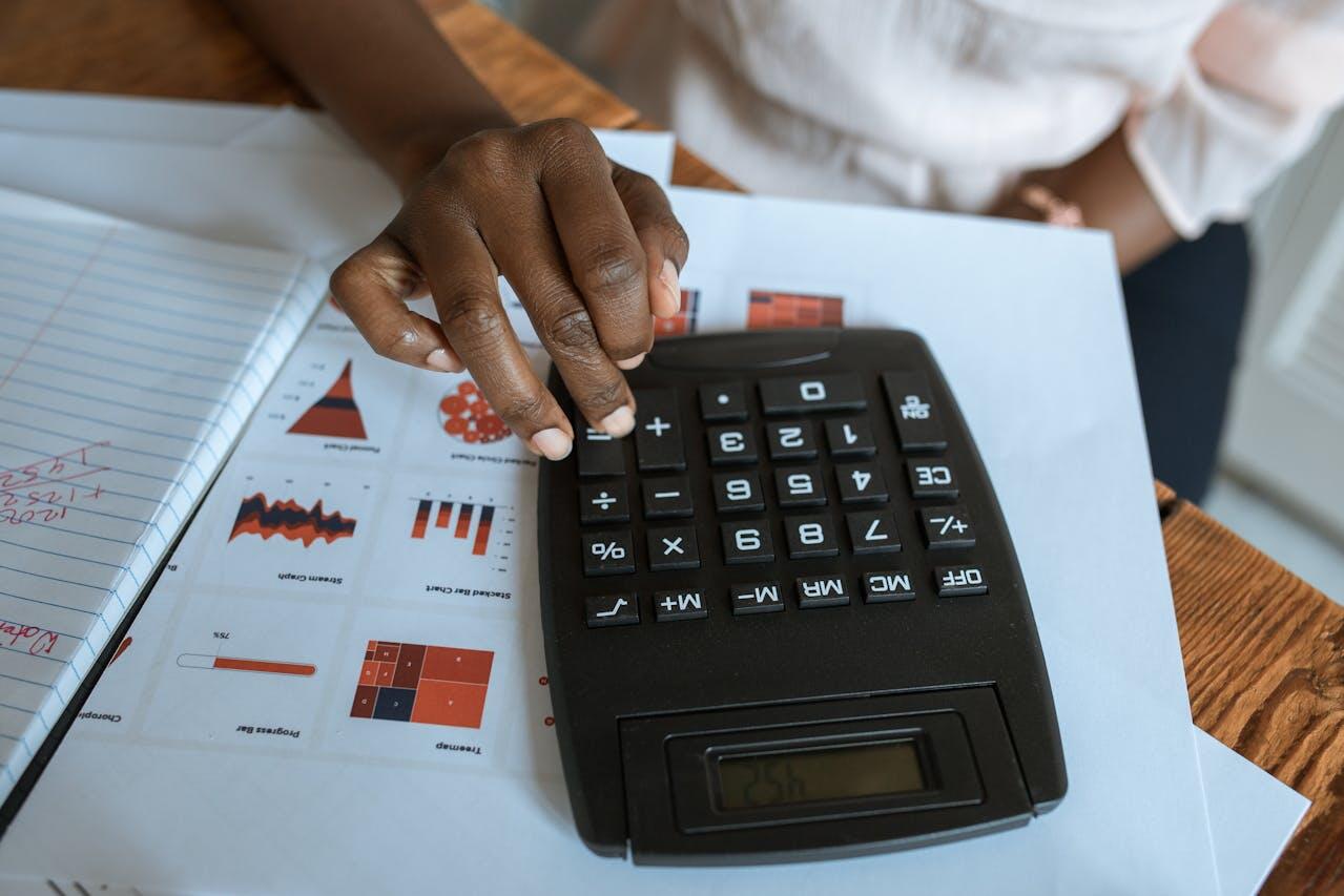 A close-up of a hand using a calculator on a desk covered with charts, graphs, and a notebook with handwritten notes.