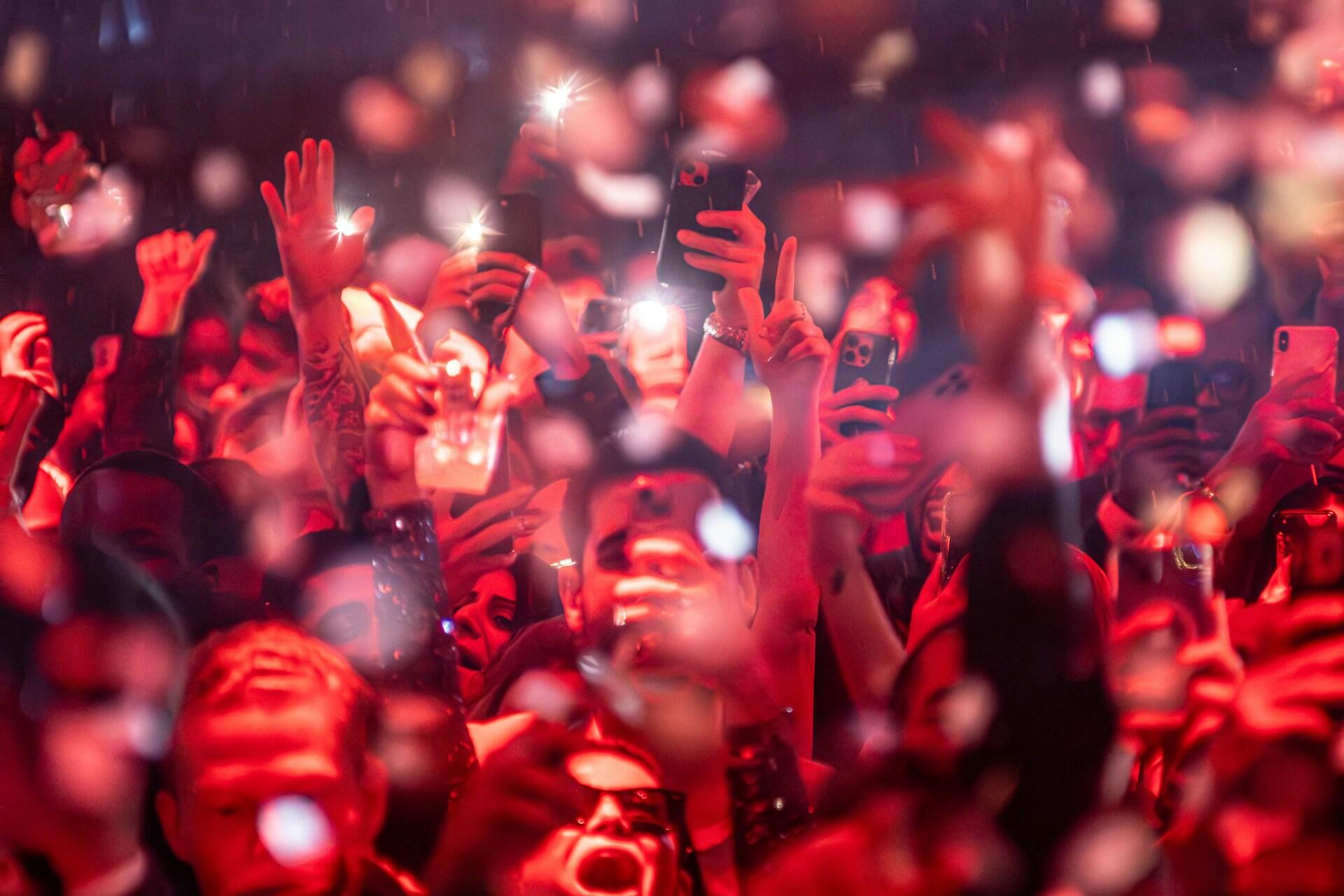 A lively concert crowd is illuminated by red lights, with many Raptors fans holding up their phones to capture the moment amidst confetti.
