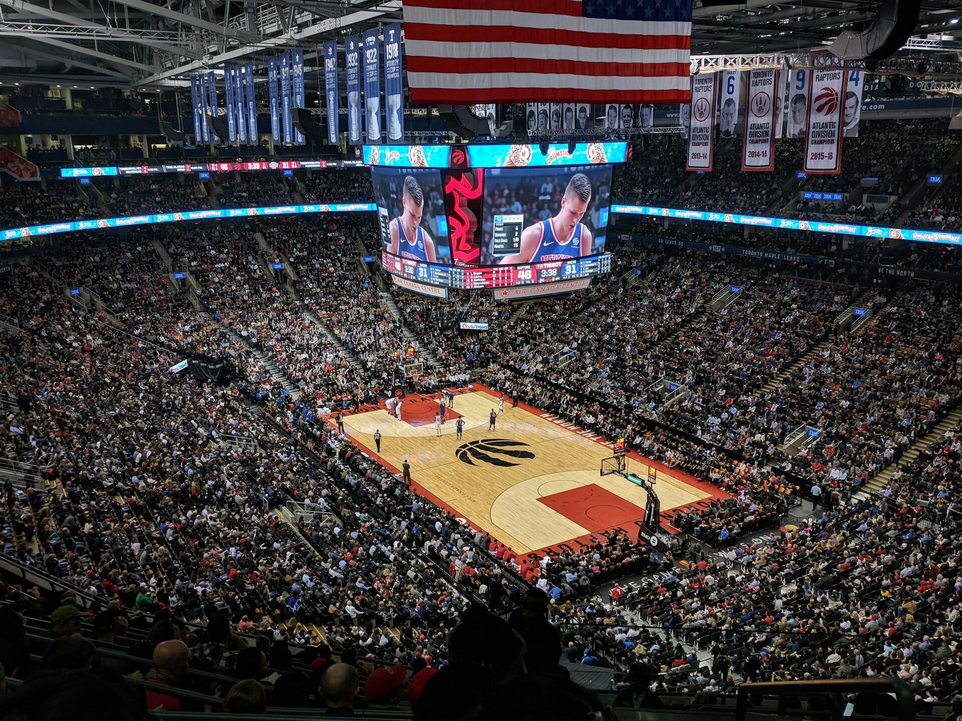 A packed basketball arena filled with fans, large screens displaying the game, and colorful team banners hanging from the rafters.