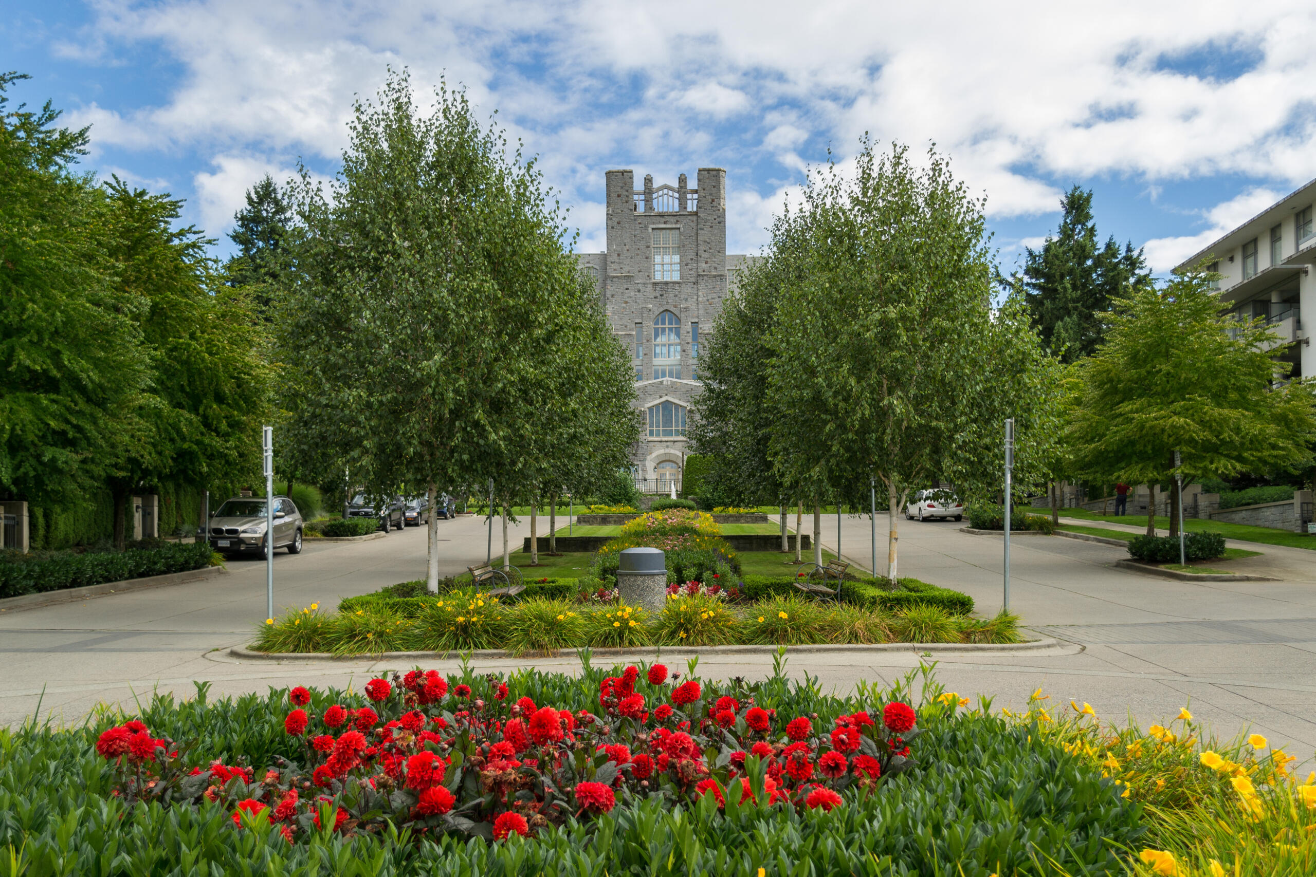 View of the University of British Columbia (UBC) campus with a tree-lined road and colorful flower beds leading to a historic stone building.