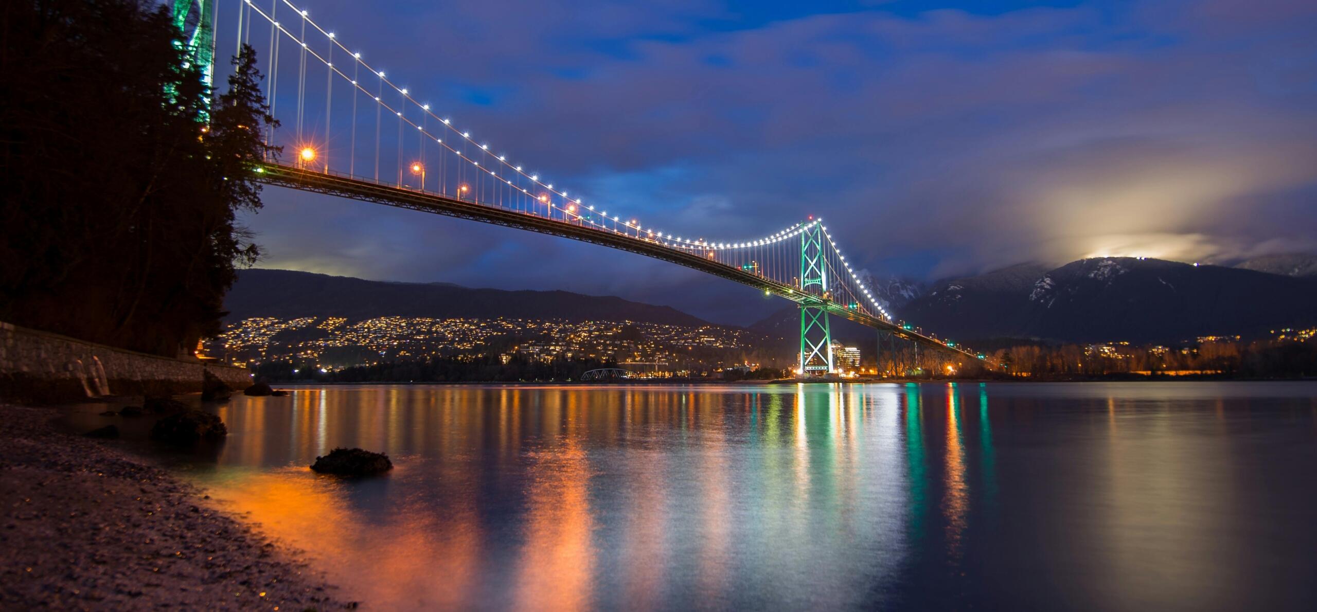 A view of a brightly lit bridge in Vancouver at night, with reflections shimmering on the water below.