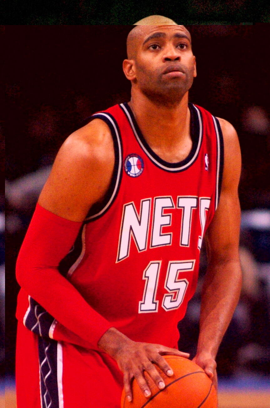 A basketball player wearing a red New Jersey Nets jersey with the number 15 prepares to shoot a basketball on the court.