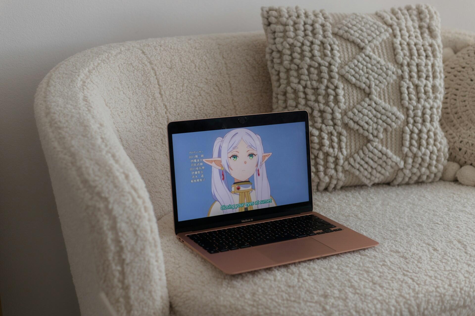 A laptop on a cozy, textured couch displays a character with long hair and large ears, with subtitles in an elegant font beneath.