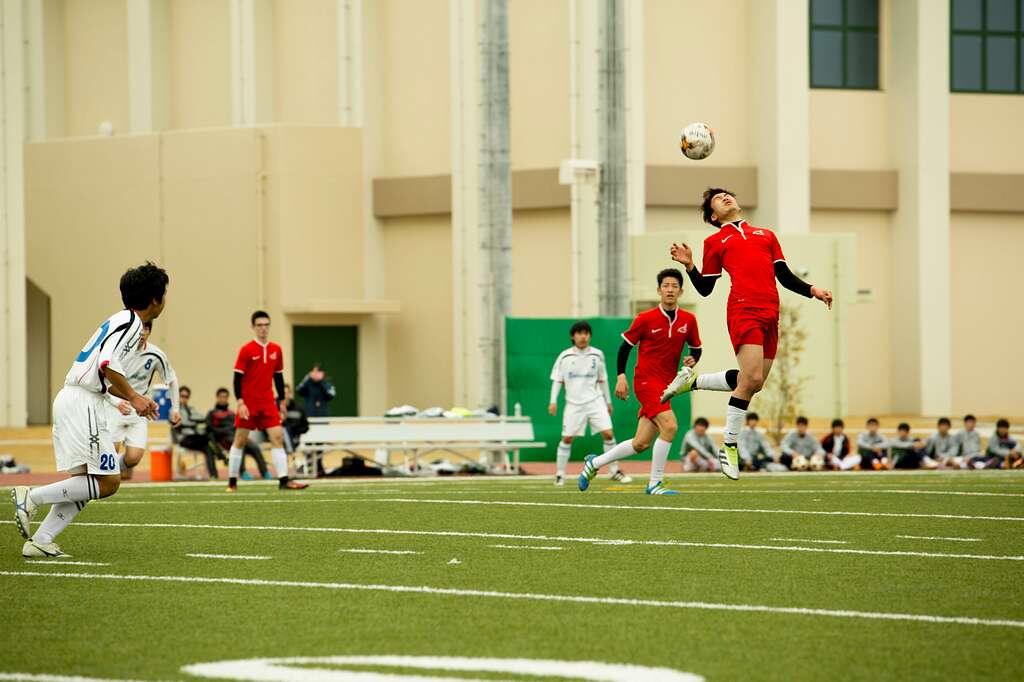  Youth soccer players in red and white uniforms competing on an artificial turf field, with one player jumping to head the ball during an academy match.