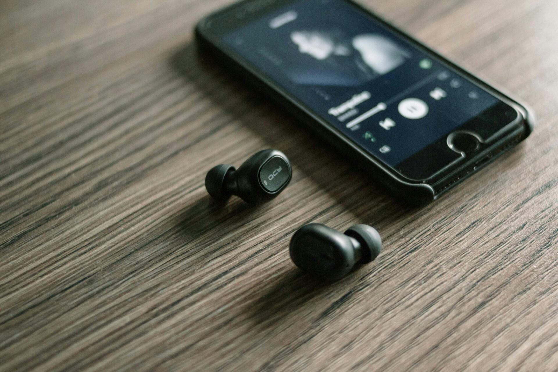 Black wireless earbuds beside a smartphone displaying a Spotify music player on a wooden surface.