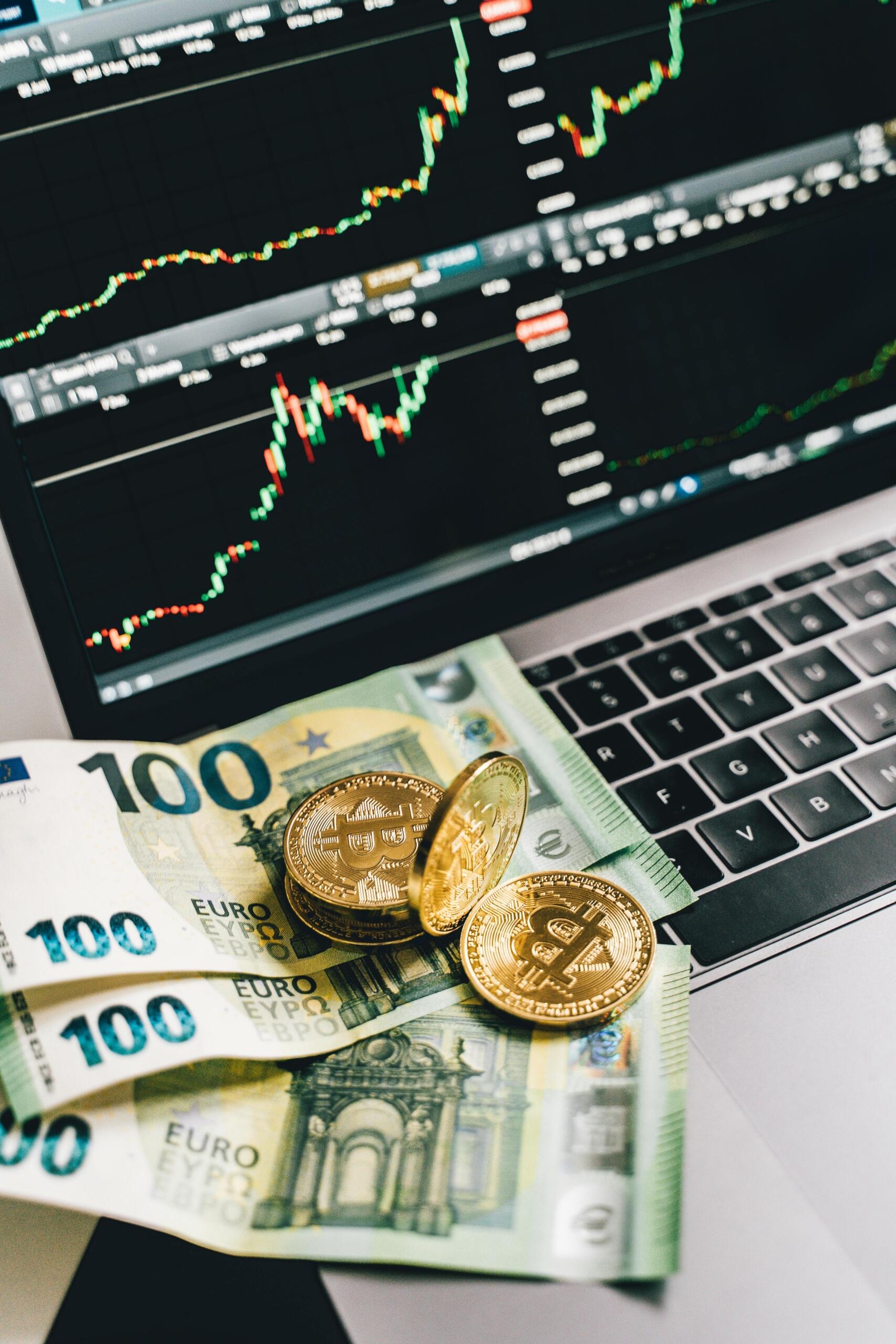 A computer screen displaying trading data and charts, with euro banknotes and Bitcoin tokens placed beside the keyboard, symbolizing global and digital currencies.