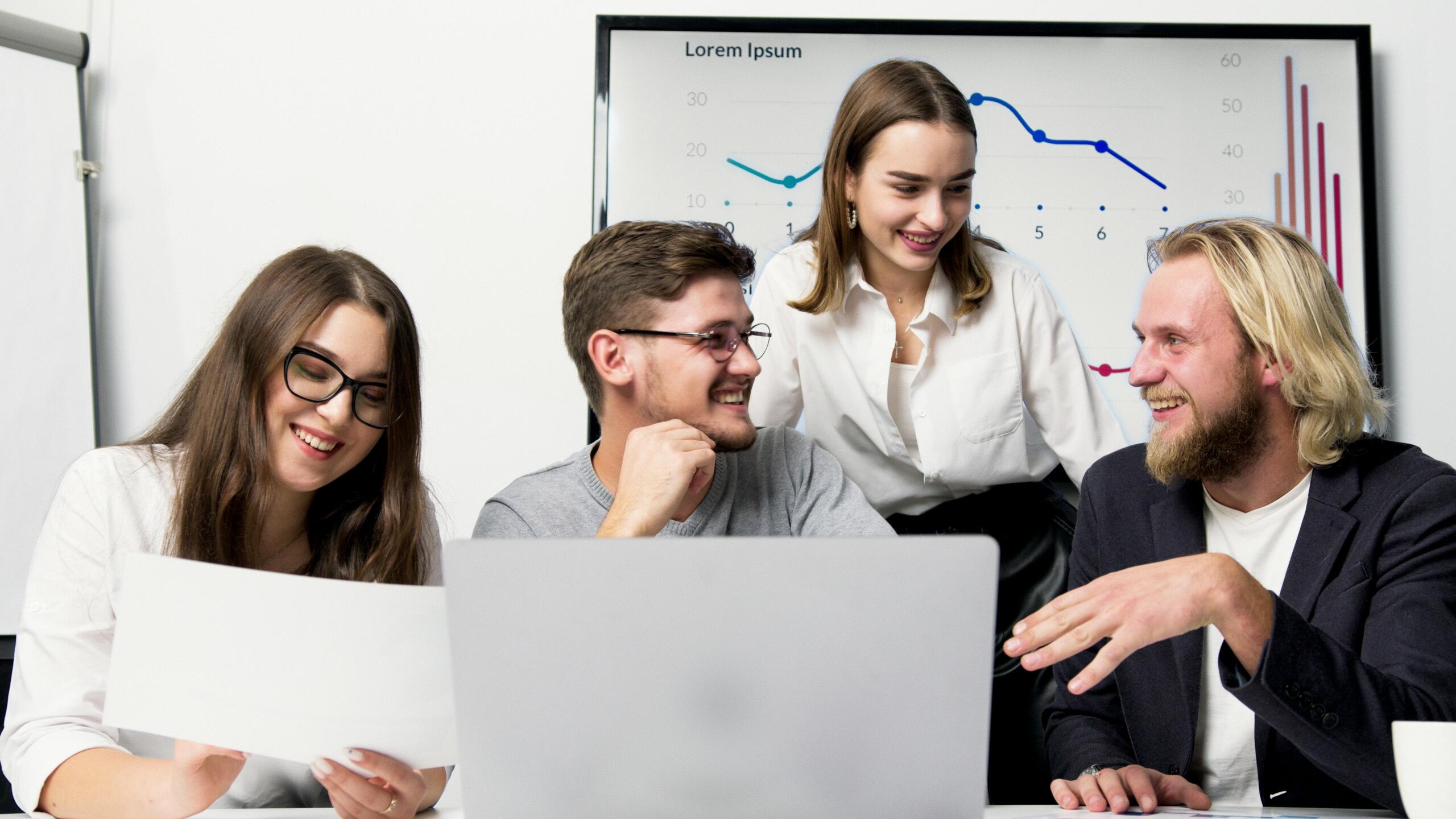 Four young professionals sitting together in front of a laptop, engaged in discussion, with a large chart displaying financial or business data behind them.