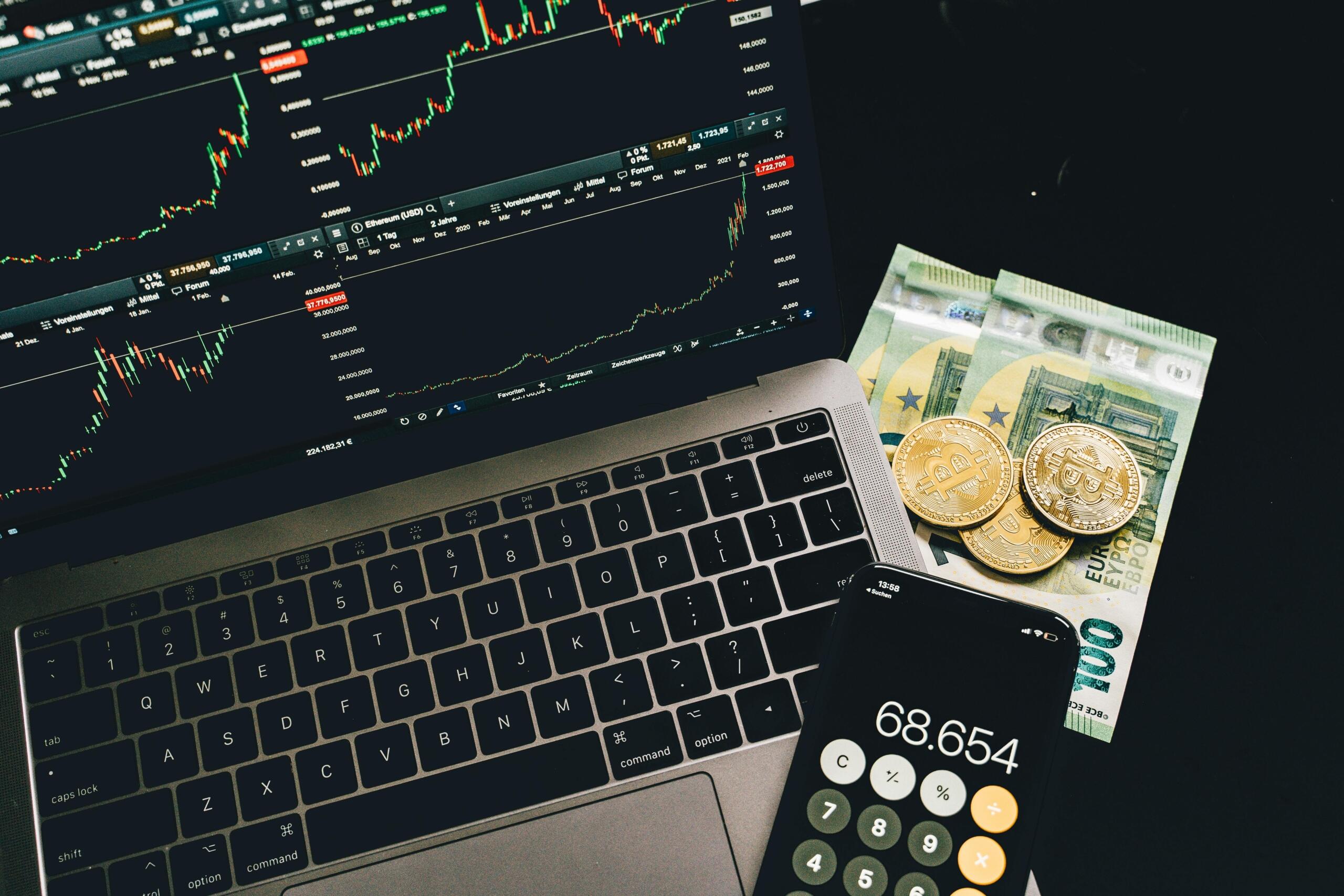 A laptop and smartphone displaying stock trading data, with scattered foreign currency notes and cryptocurrency coins beside them, representing global and digital finance.