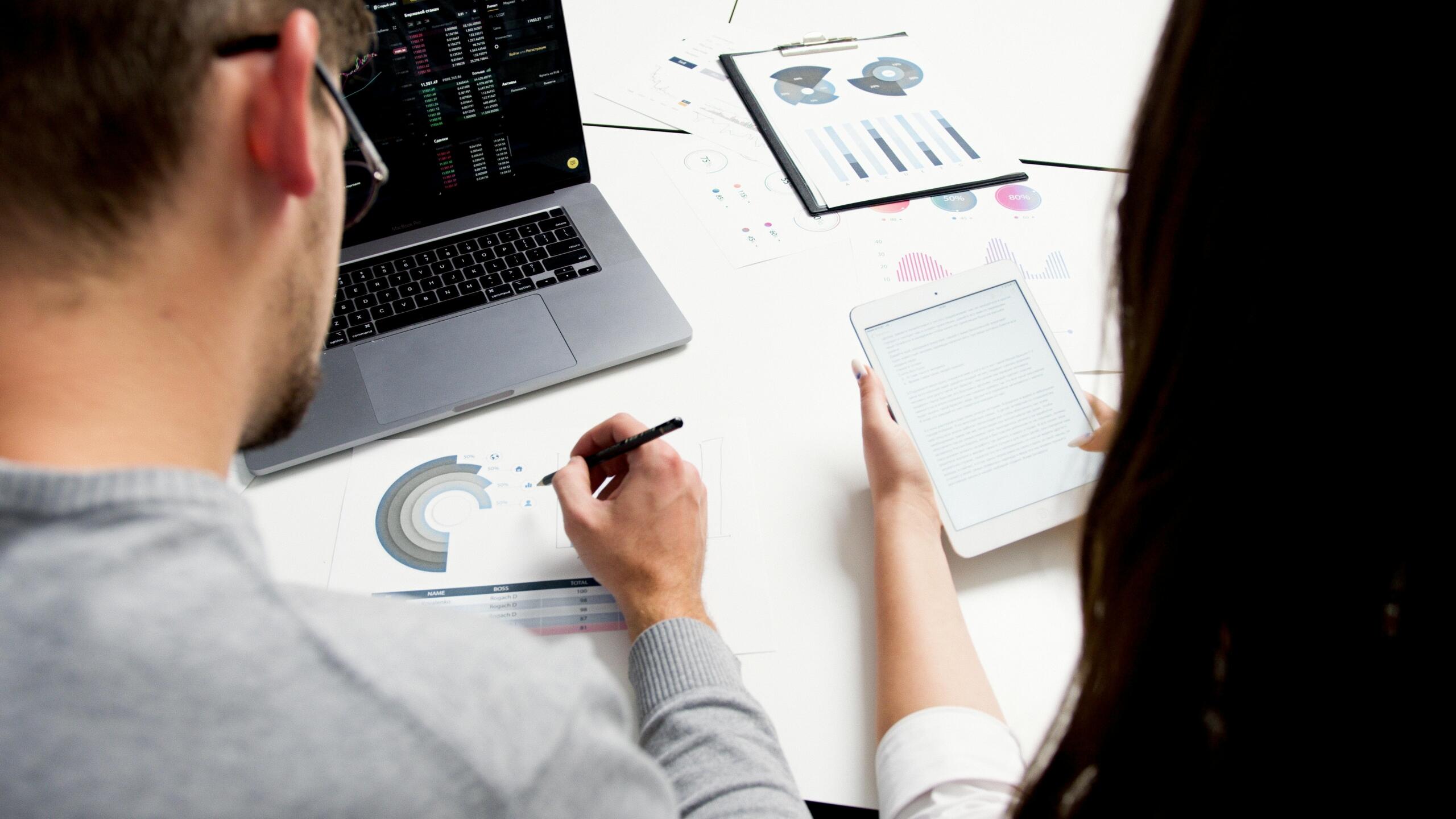A man and woman sitting side by side in front of a laptop displaying trading data, both focused on analyzing information.