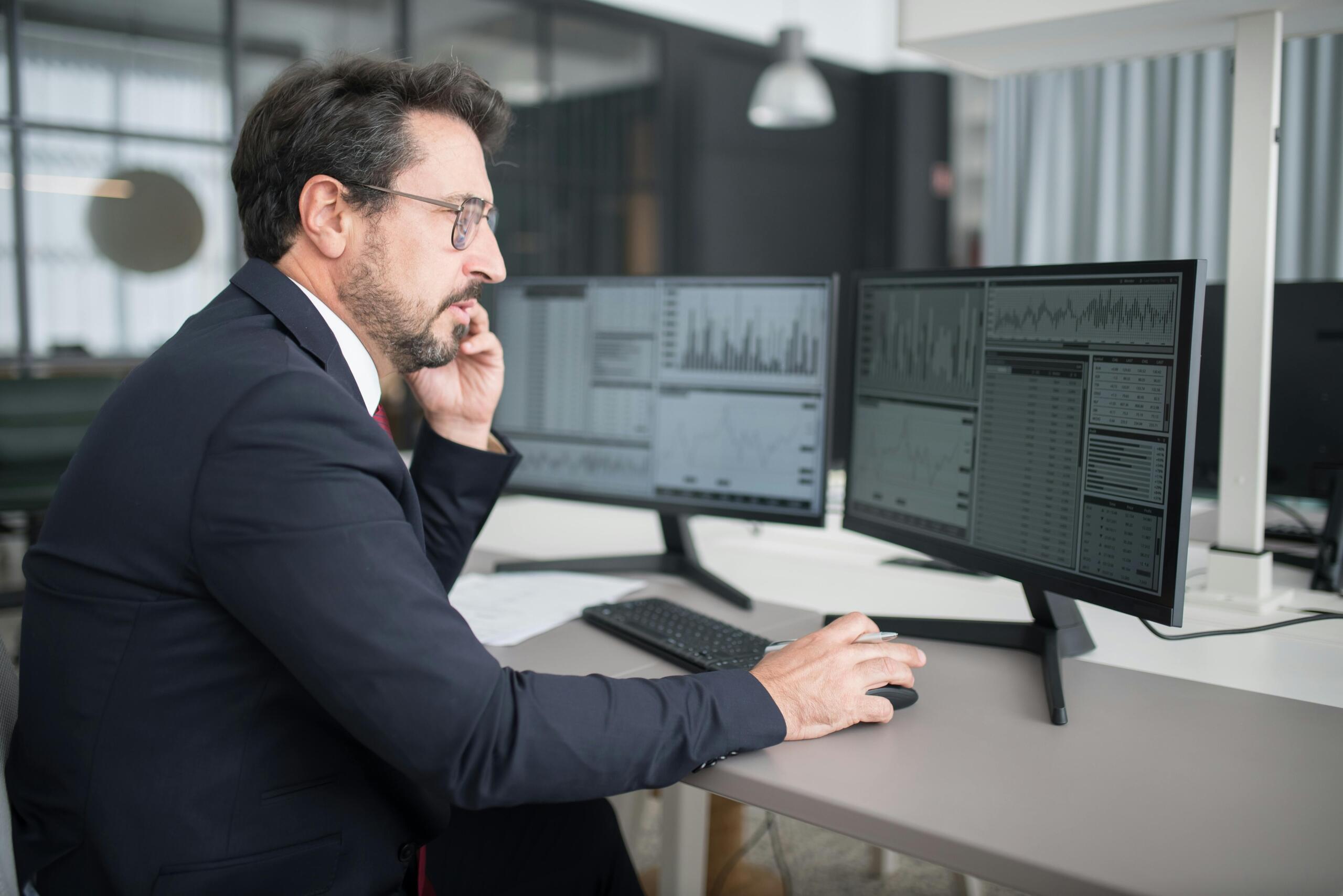 A man in a suit intently looking at trading data on a computer screen, analyzing charts and market trends in a professional setting.
