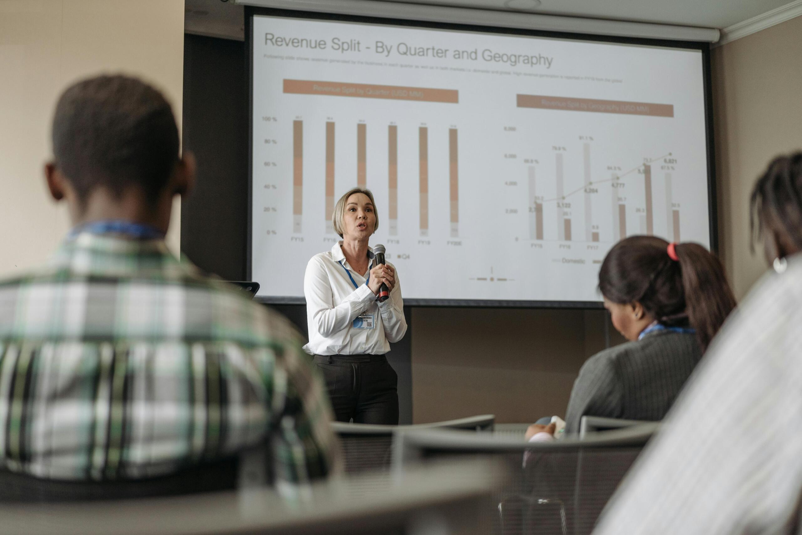 A woman holding a microphone, confidently presenting business information to an audience, with charts and data visible on a screen behind her.