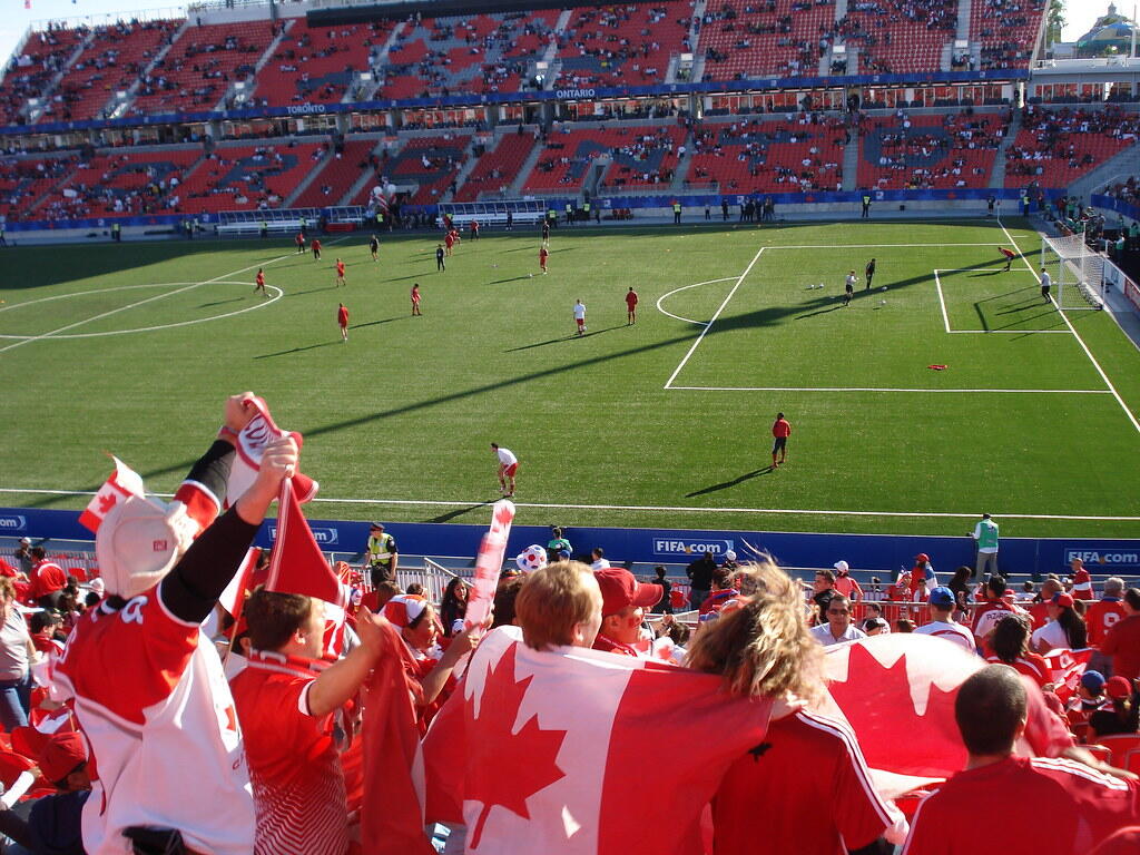 Canadian soccer fans waving flags and cheering in a stadium during a live match, with players warming up on the field in the background.