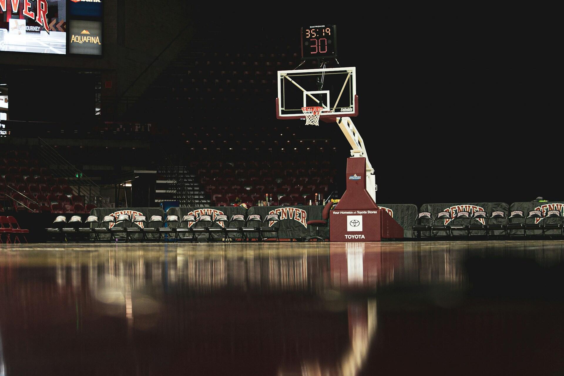 A basketball hoop and court with visible team benches in a dimly lit arena, featuring a game timer and scoreboard.