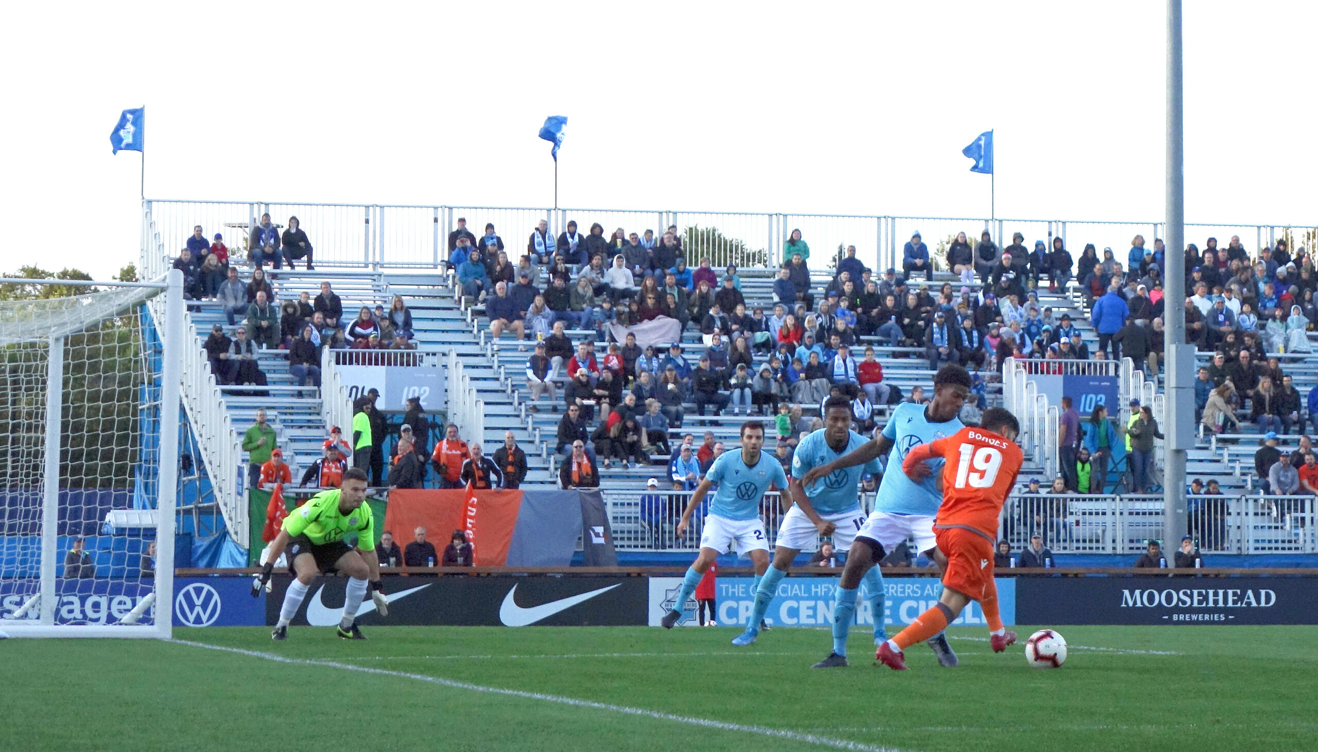 Canadian Premier League match in action, with a player in an orange jersey taking a shot on goal while defenders and a goalkeeper prepare to block, in front of a full stadium crowd.