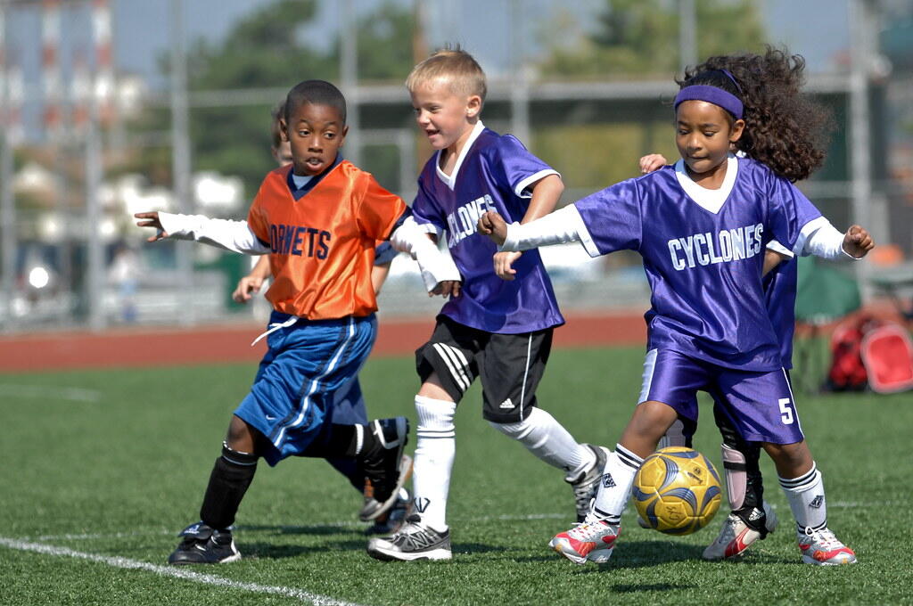 Young children from two soccer teams, the Hornets and Cyclones, compete for the ball during a lively youth soccer match on a sunny day.