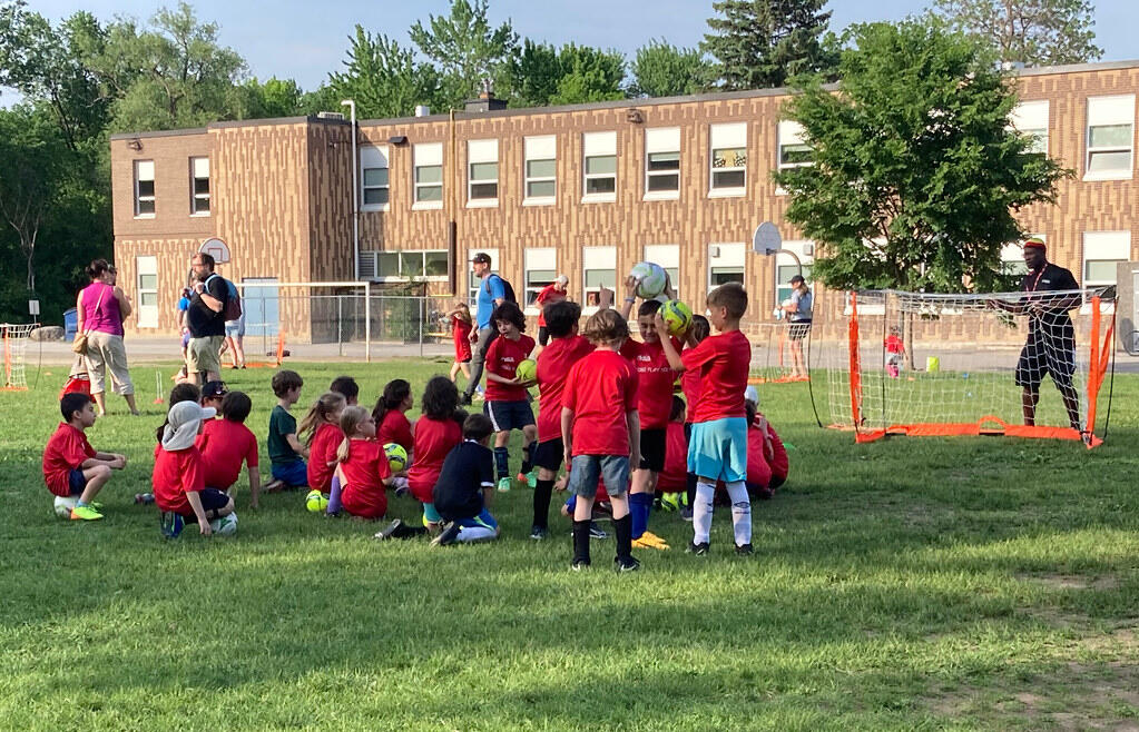 Children participating in a community soccer program on a grassy field outside a school, with coaches and small goals set up for training.