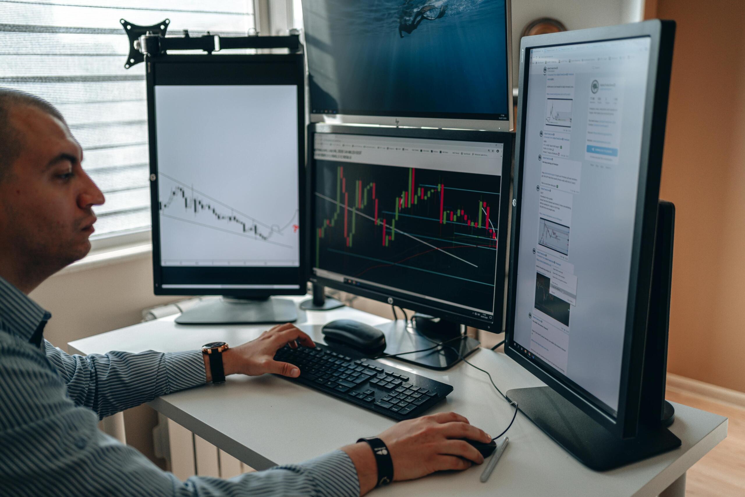A man trading stocks at his desk, focused on two computer screens displaying financial charts, stock prices, and market data.