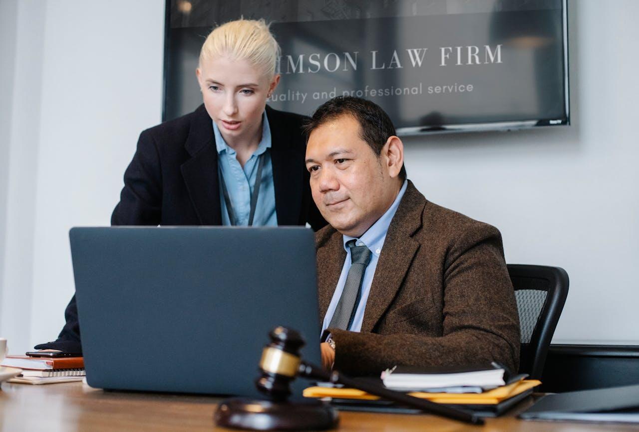 A lawyer and an assistant collaborate at a desk, discussing a laptop with legal documents and a gavel in view.