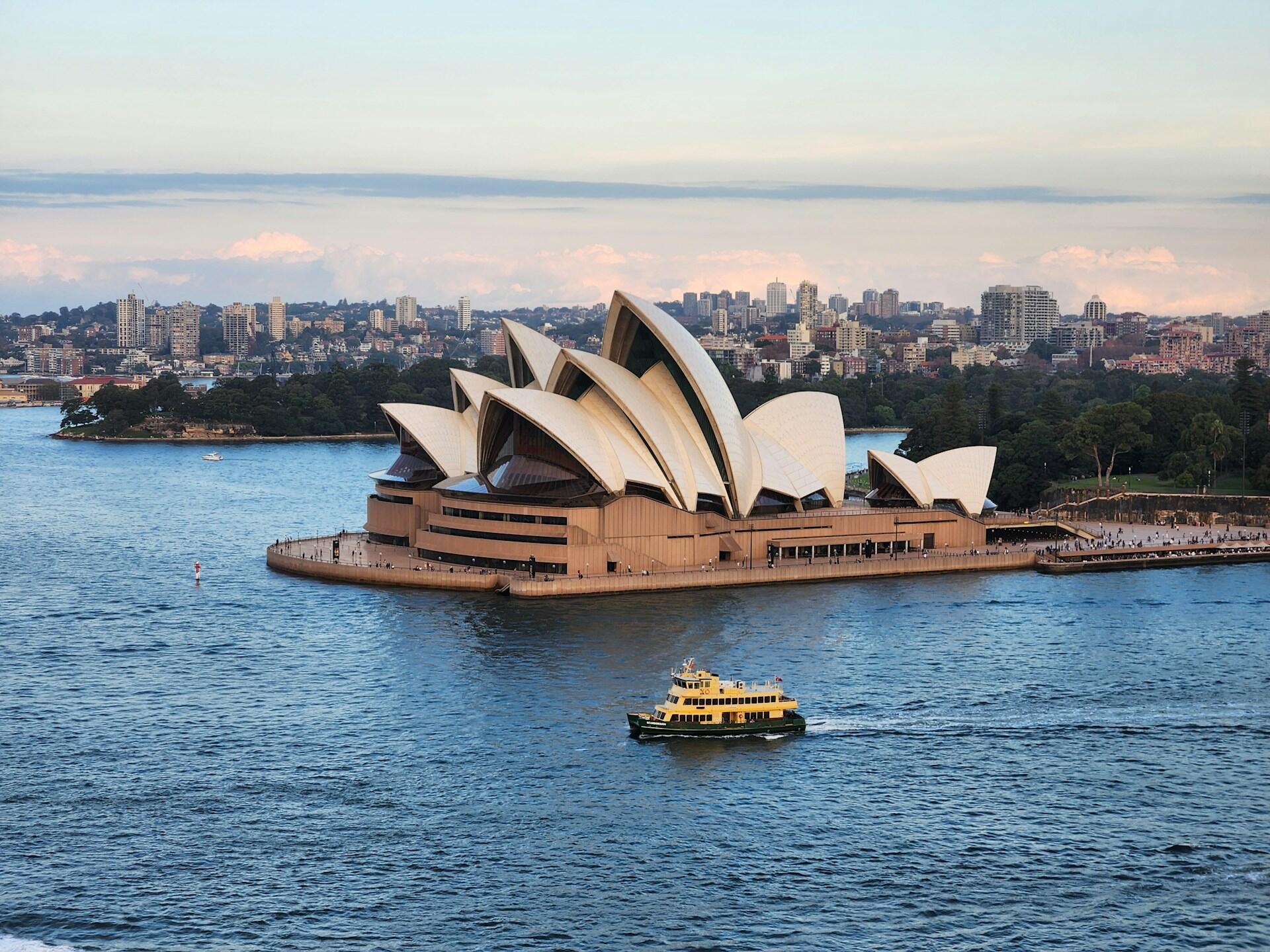 Sydney Opera House with its iconic sail-like design, a ferry navigating the blue waters, and the city skyline in the background.