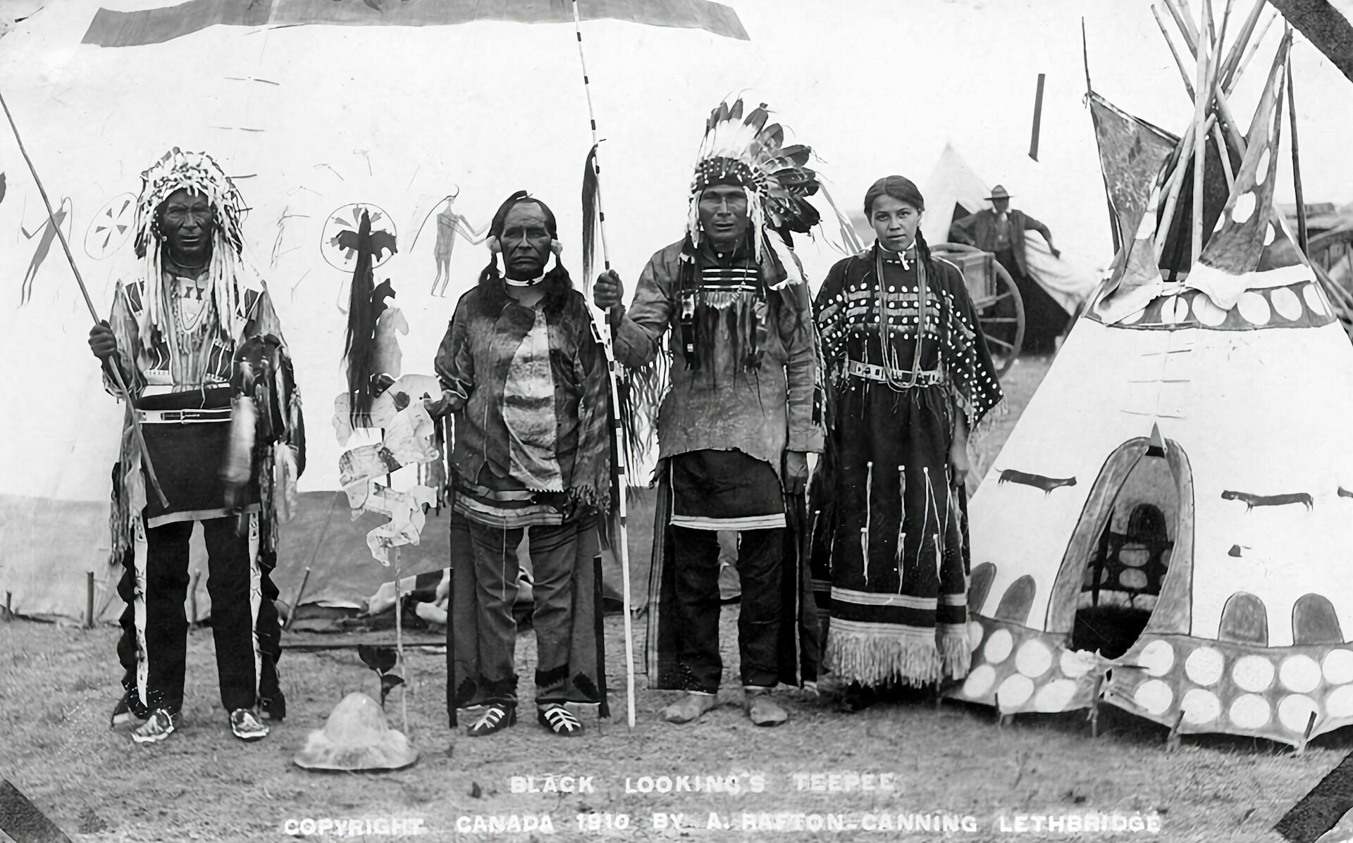Four Indigenous individuals stand in front of a traditional teepee, dressed in ceremonial attire with intricate patterns and accessories.