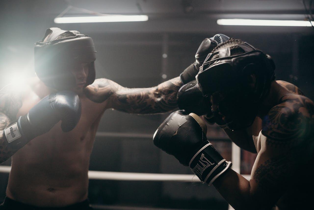 Two tattooed boxers spar in a ring, wearing protective gear and focused expressions, with a dramatic lighting effect in the background.
