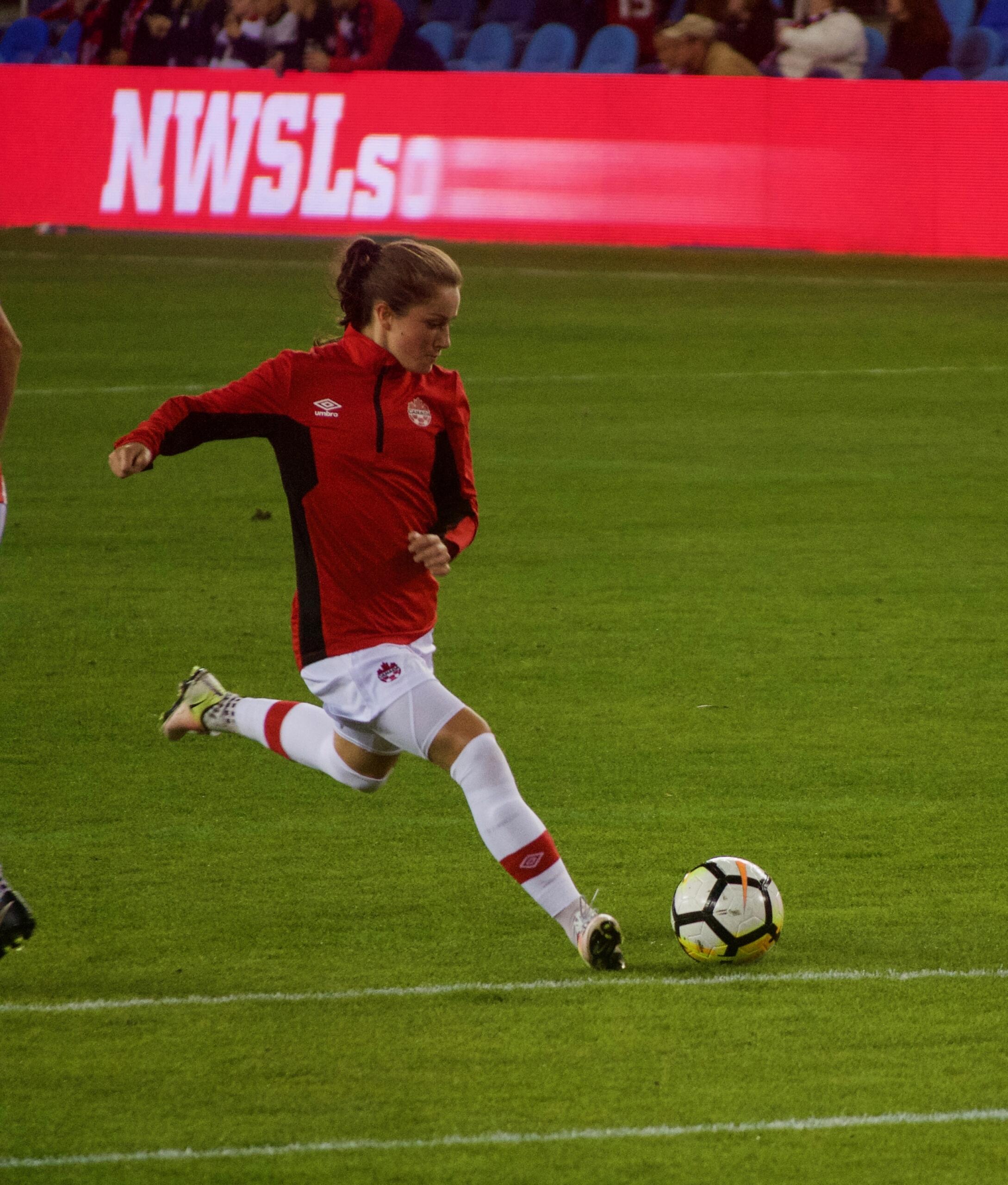 Canadian soccer player Jessie Fleming takes a powerful shot during warm-up, wearing a red national team jacket on a green field with “NWSL” signage in the background.