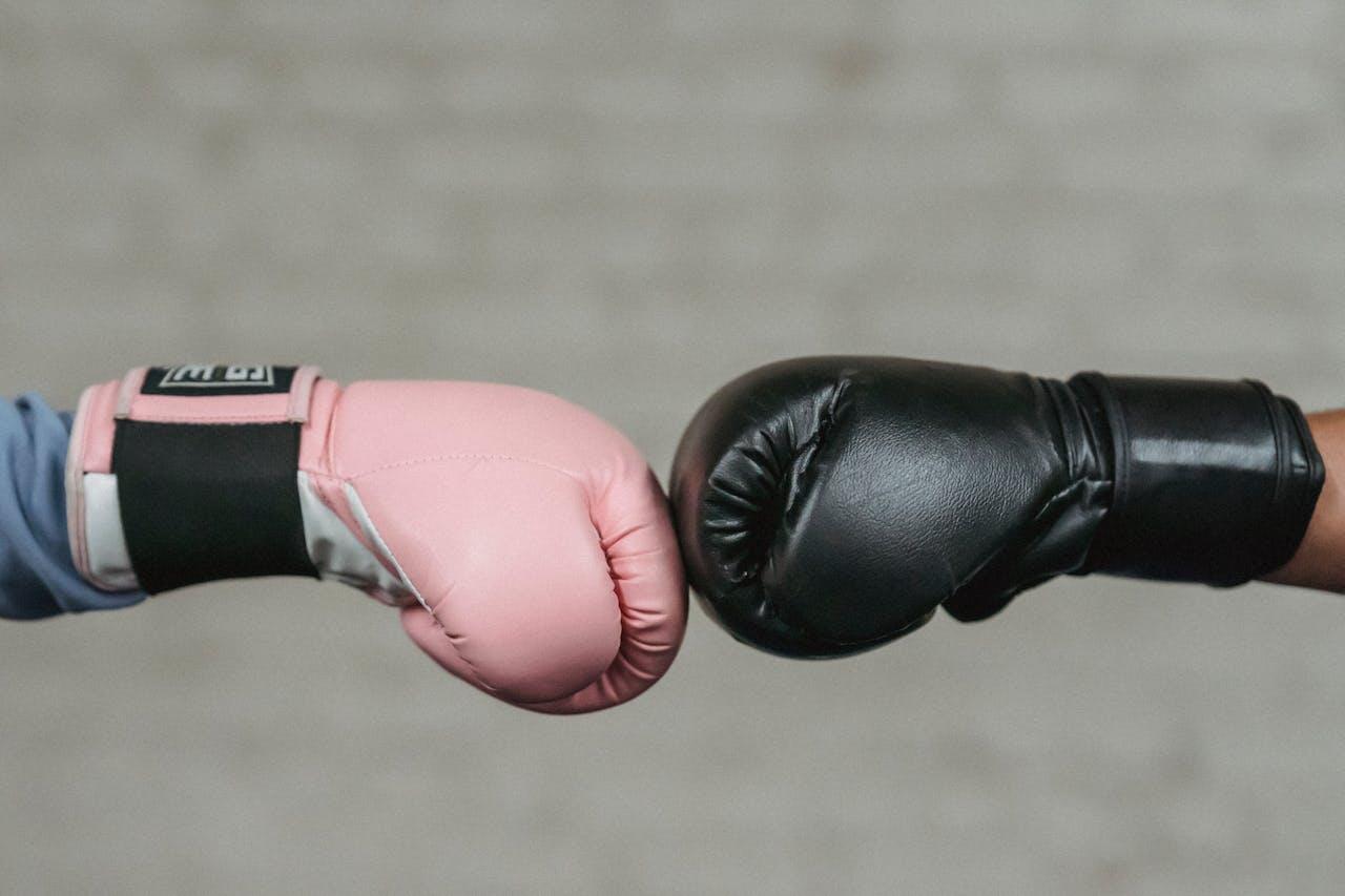 Two boxing gloves, one pink and one black, meet in a fist bump against a textured gray background.