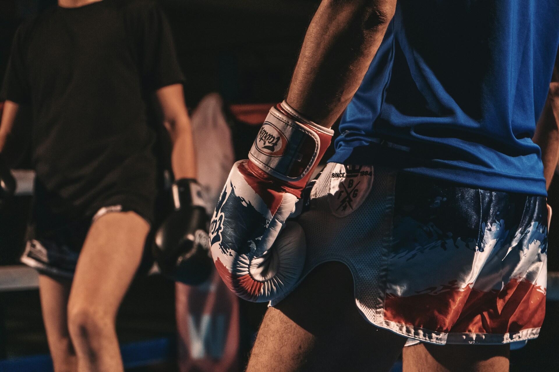 Two boxers in a ring: one in a blue shirt and colorful shorts holds red and white gloves; the other, in black, prepares for the match.