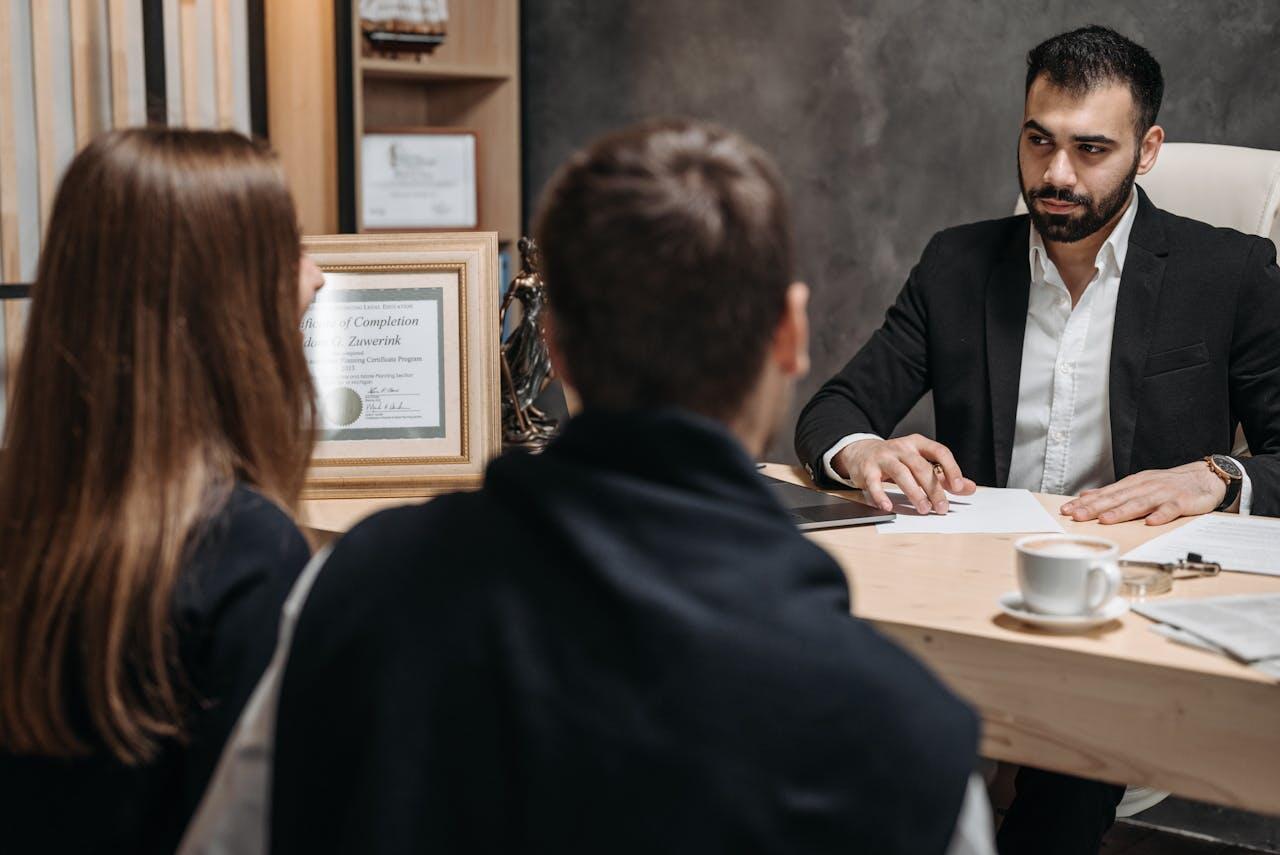 A man in a suit discusses paperwork with a couple in an office, featuring a certificate and a cup of coffee on the table.