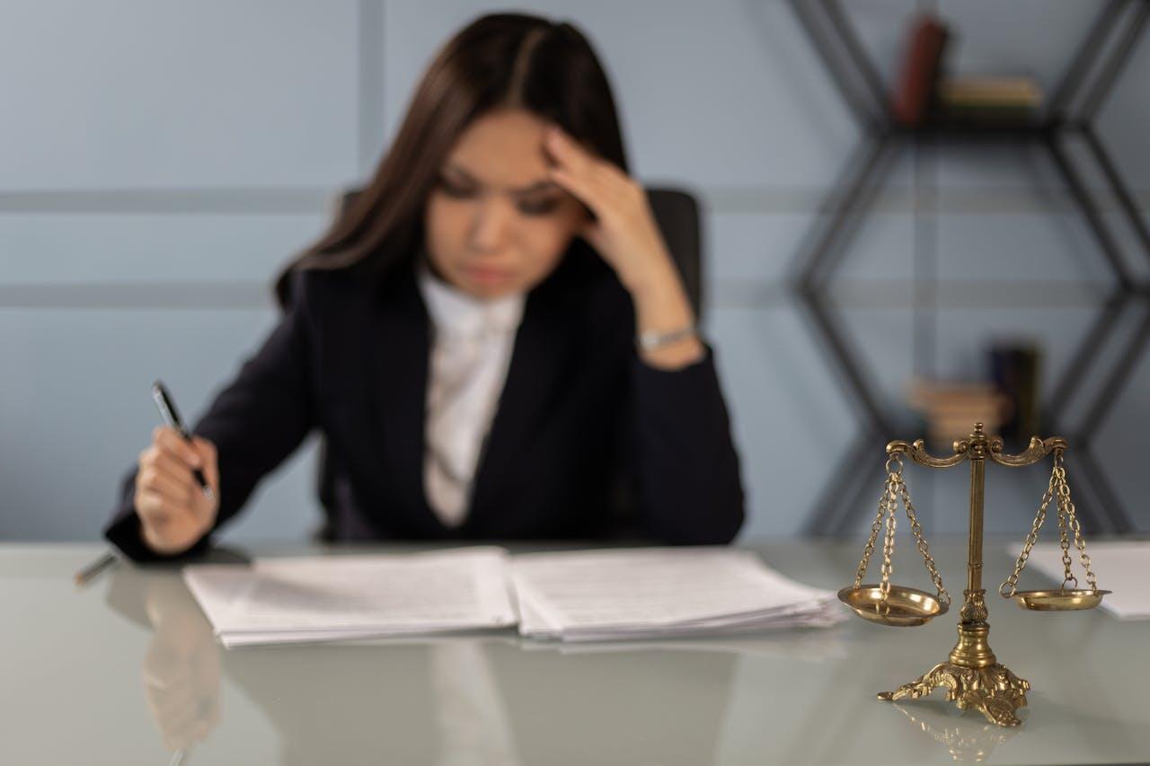 A professional woman in a dark suit sits at a desk with papers, holding a pen, while a decorative scale of justice is nearby.
