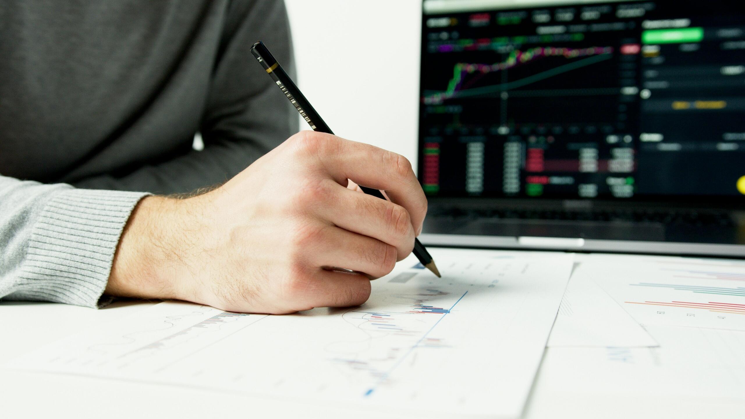 A computer screen displaying stock market data while a man's hands are seen in the foreground writing notes on paper.