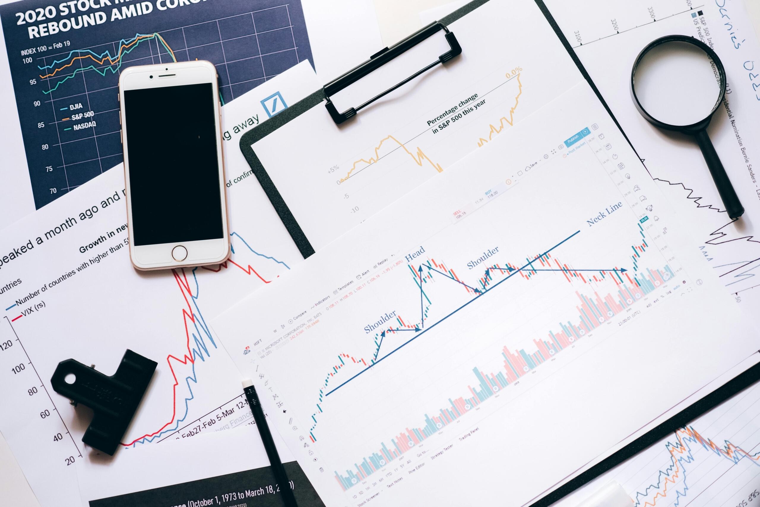 A table covered with printed stock market graphs, a digital tablet displaying financial data, and a magnifying glass placed on top, suggesting detailed market analysis.