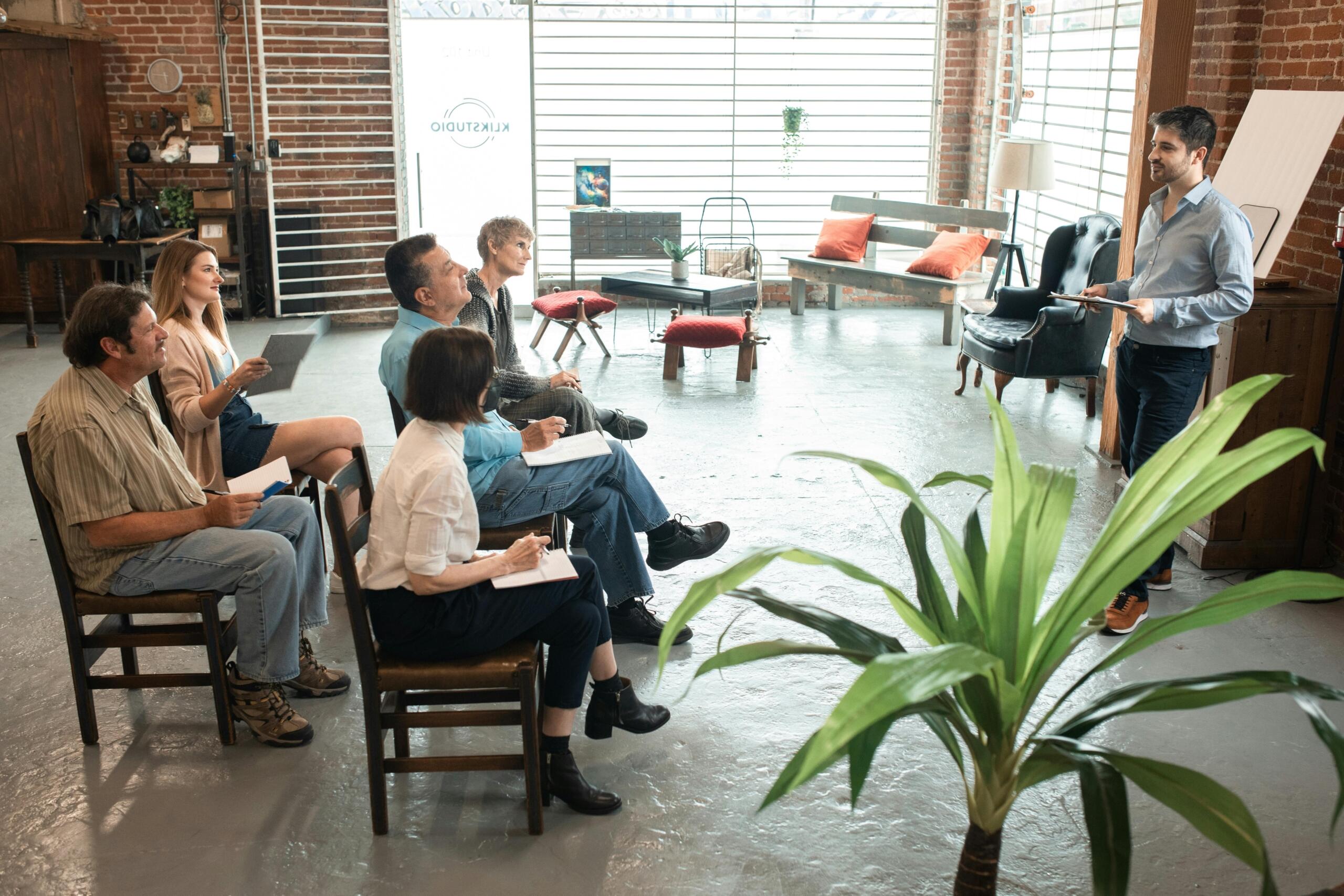 A man standing at the front of a classroom presenting a workshop to a group of attentive adult learners seated at tables, with a whiteboard and presentation screen behind him.A man presenting a workshop to a group of attentive adult learners seated in chairs. The attendees take notes.
