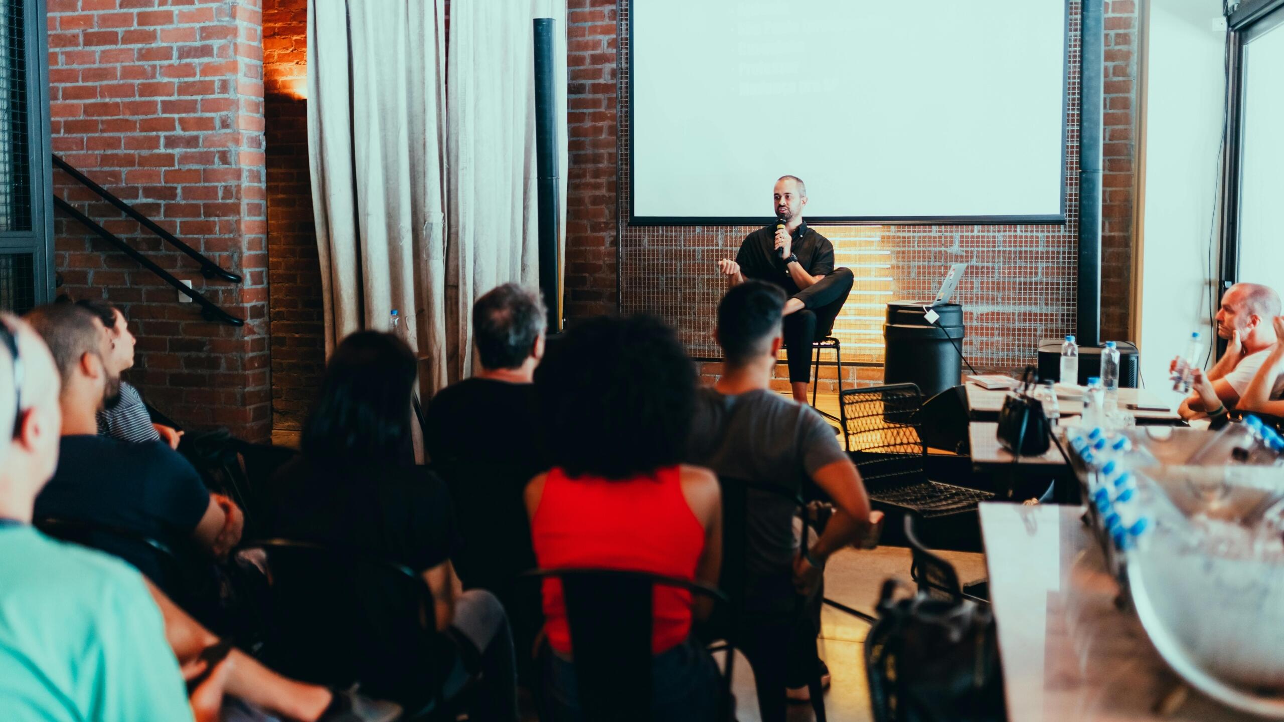 A man delivering a workshop on a stage with exposed brick walls, speaking to a seated audience in a casual, professional setting.