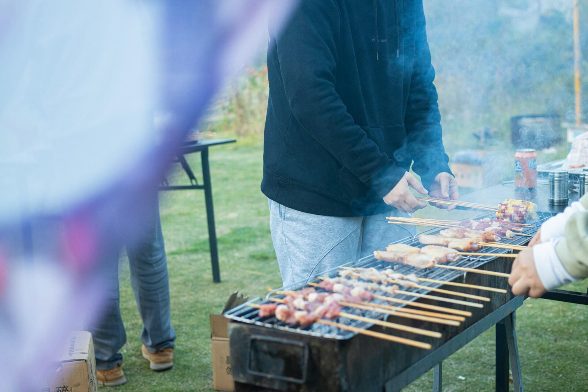A person in a black hoodie grills skewers over a smoky surface, surrounded by green grass and other people in the background.